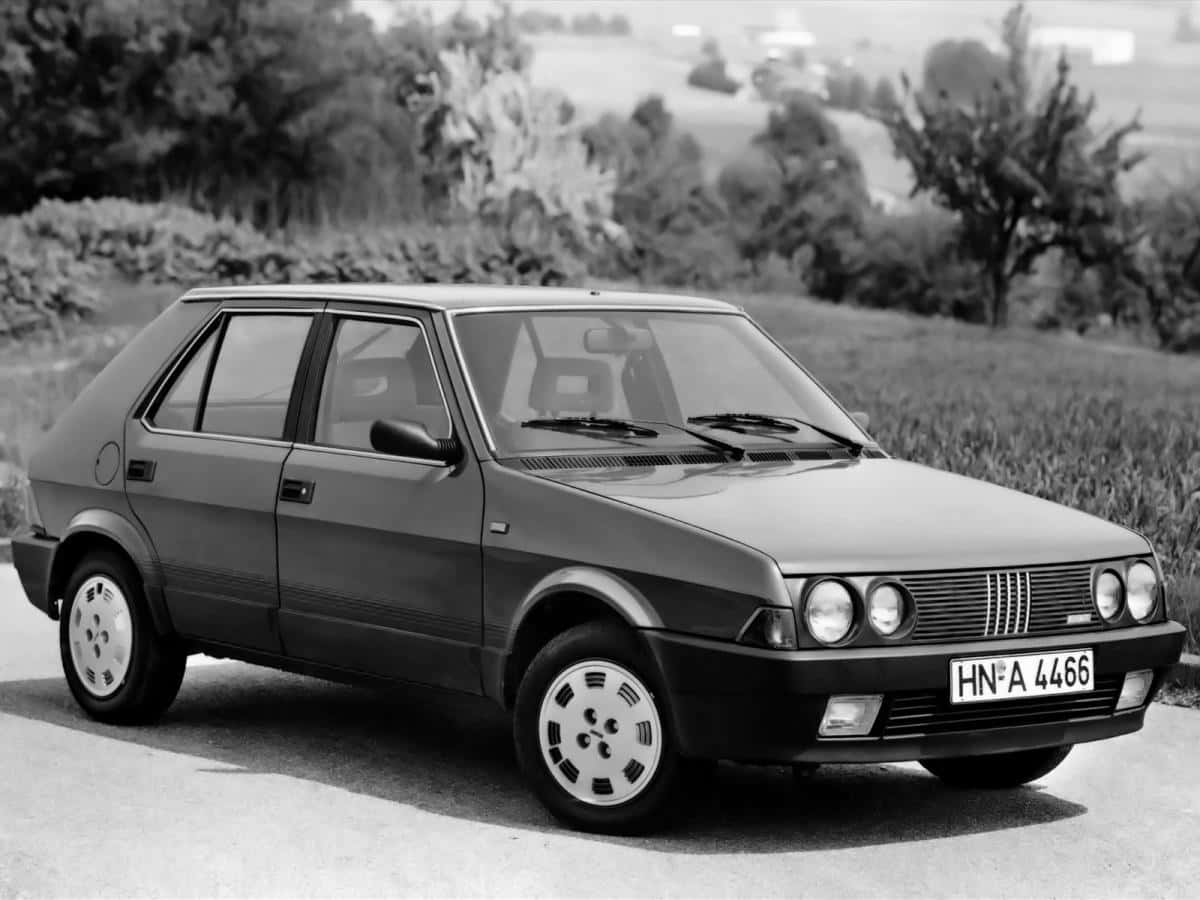 Fiat Ritmo 桌布 壁紙 — 一輛經典的Fiat汽車停泊在鄉村道路上，背景有樹木和田野，黑白風格。