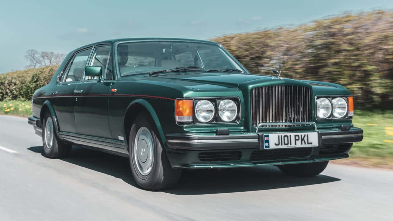  壁紙 — 一輛經典綠色 Bentley Turbo R 在陽光明媚的鄉村道路上巡航，背景是模糊的樹籬。