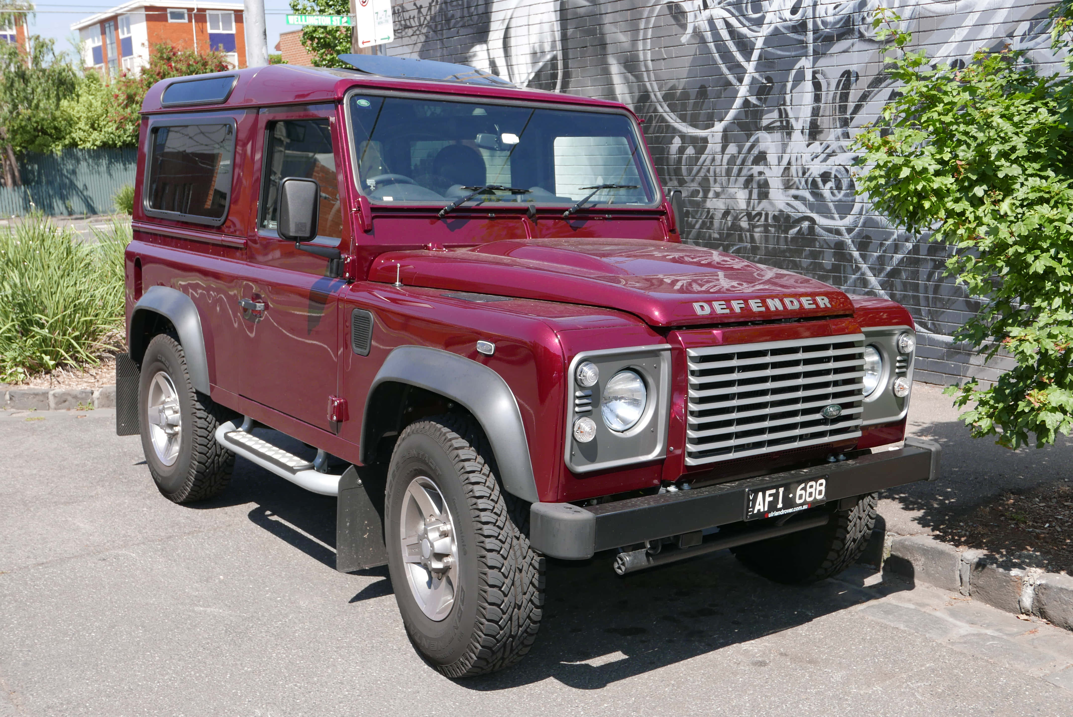 Land Rover Defender 桌布 壁紙 — 一輛經典的栗色Land Rover Defender停泊在陽光明媚的街道上，旁邊是佈滿塗鴉的牆壁和綠植。