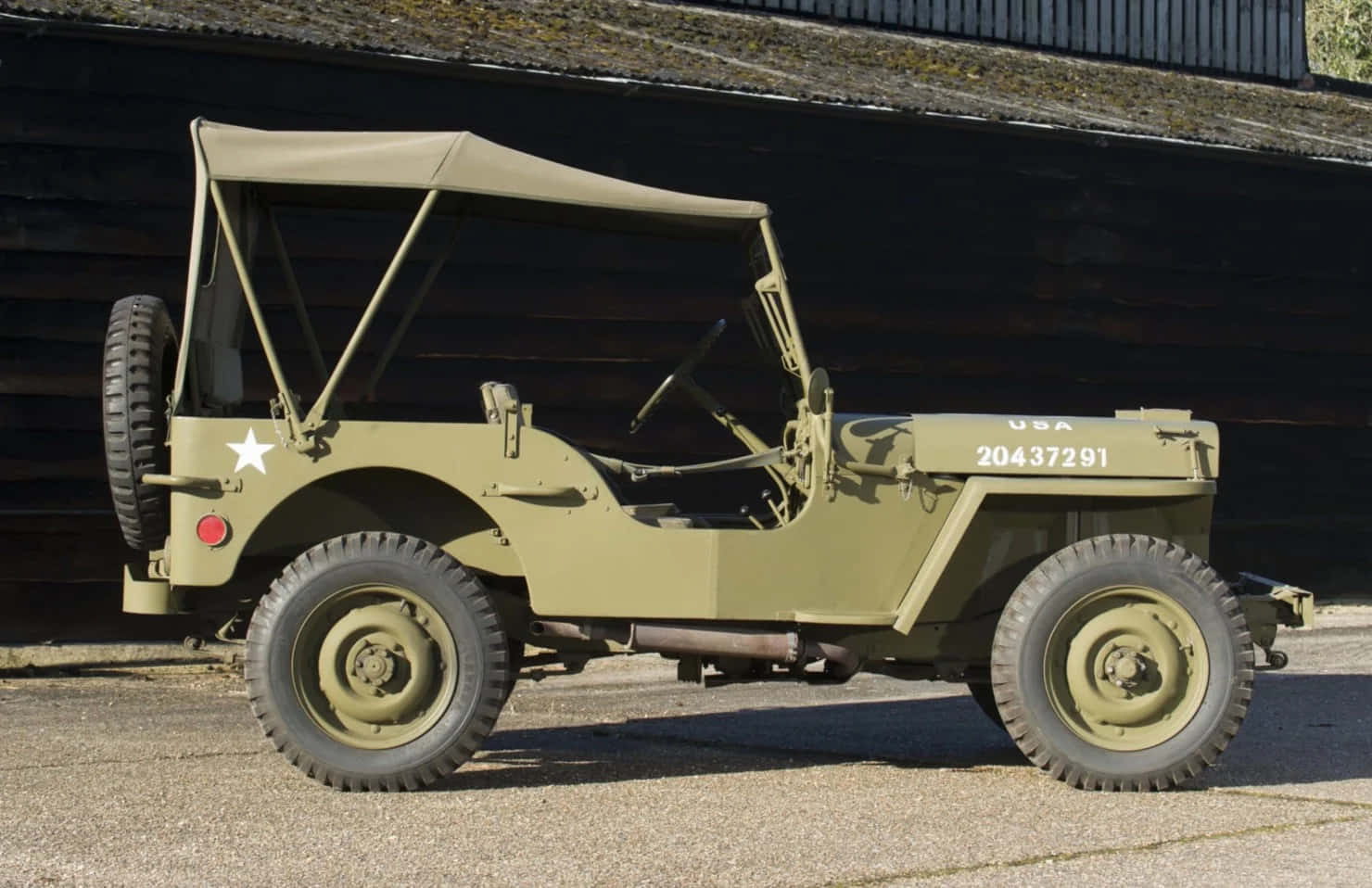 Jeep Willys 桌布 壁紙 — 一輛經典橄欖綠軍用吉普車，帶有白色星星標誌，停在碎石上，旁邊是深色木牆，在明亮的陽光下。