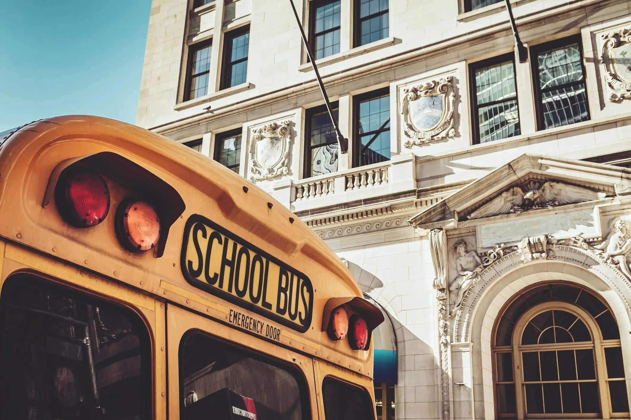 School Aesthetic 桌布 壁紙 — 一輛經典黃色校車停泊在裝飾華麗的石建築前，天空清澈湛藍。