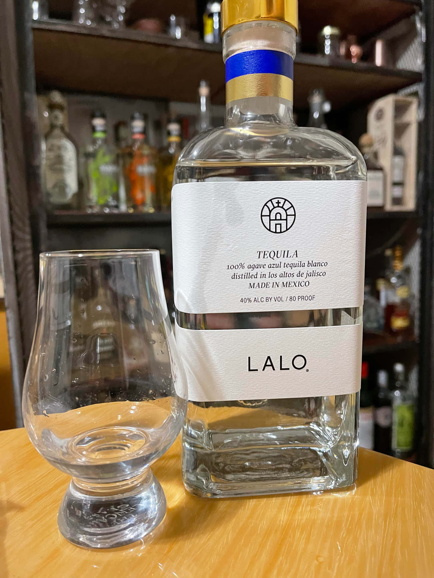  壁紙 — 一個透明嘅Lalo tequila blanco酒樽放在一個空嘅品酒杯旁邊，背景係一個有酒架嘅木吧檯。