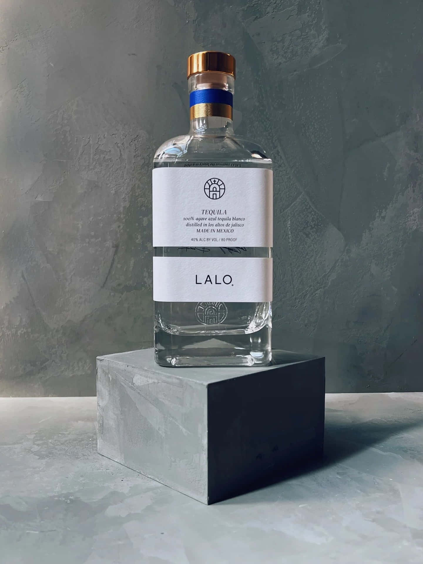  壁紙 — 一個透明嘅Lalo龍舌蘭酒樽，金色樽蓋，優雅咁擺喺灰色石座上面，背景係有紋理嘅灰色牆。