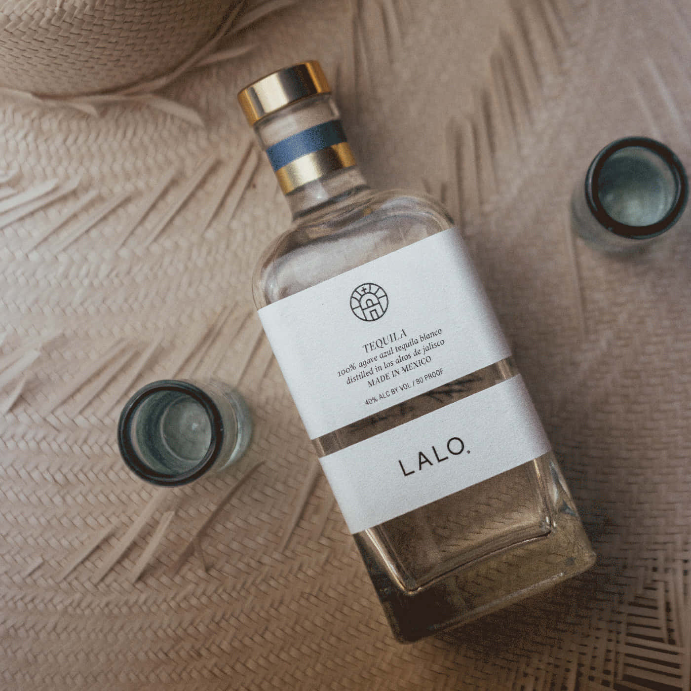  壁紙 — 一個標有 Lalo 的透明龍舌蘭酒瓶放在編織米色墊子上，旁邊有兩個小玻璃杯。