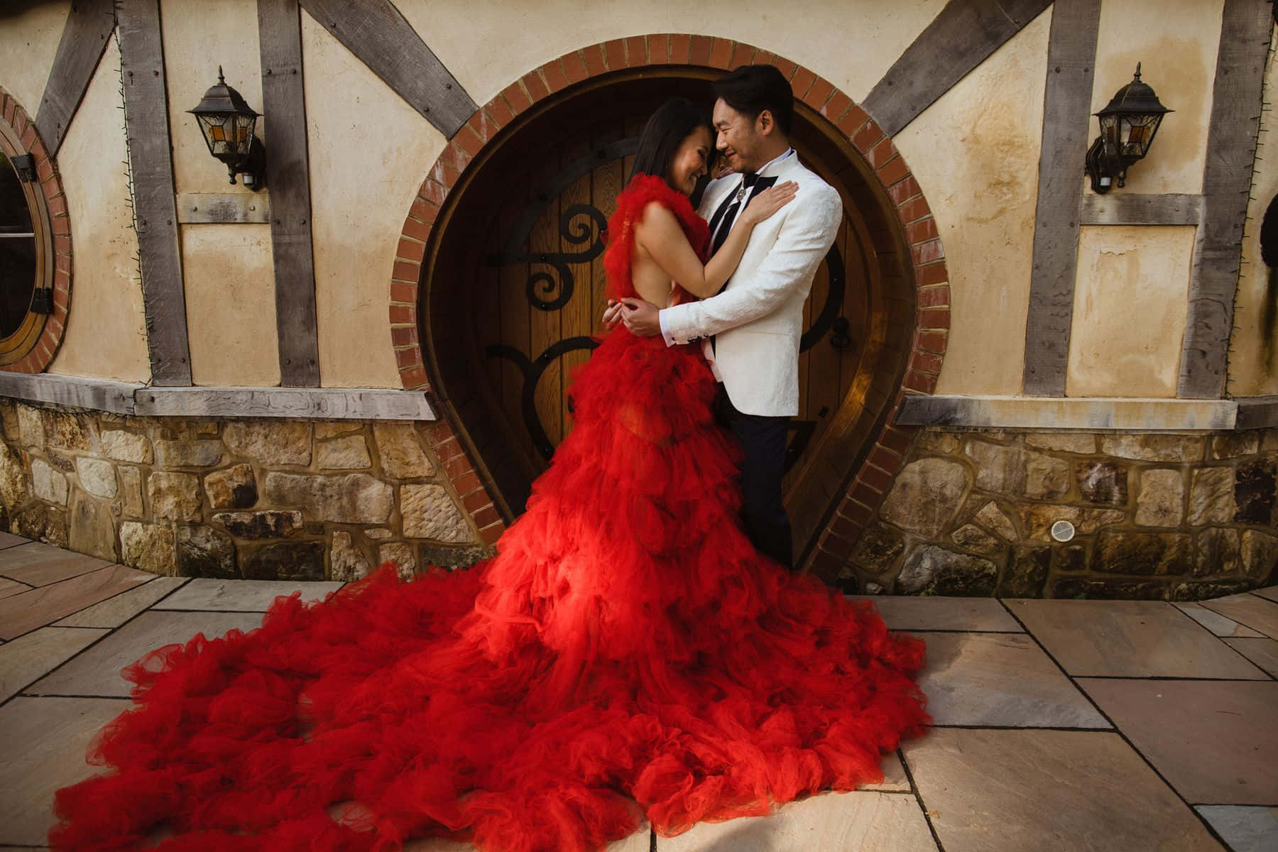 Red Wedding 桌布 壁紙 — 一對情侶在質樸的心形門前溫柔擁抱，女士穿著寬大的紅色羽毛禮服，男士穿著白色燕尾服。