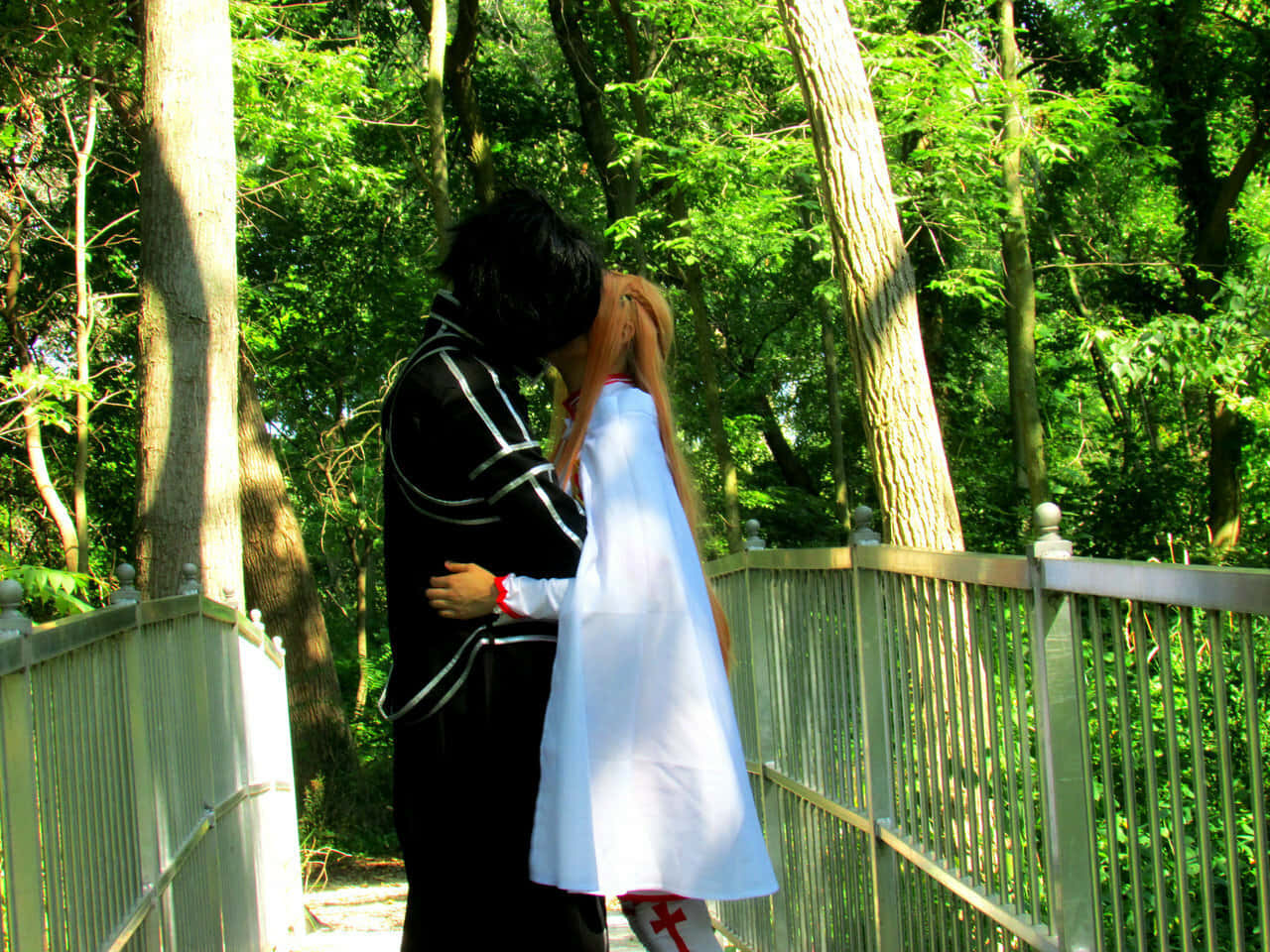 Sword Art Online Cosplay 桌布 壁紙 — 一對穿著角色扮演服裝的情侶在陽光明媚的森林橋上溫柔擁抱，周圍是茂密的綠樹。