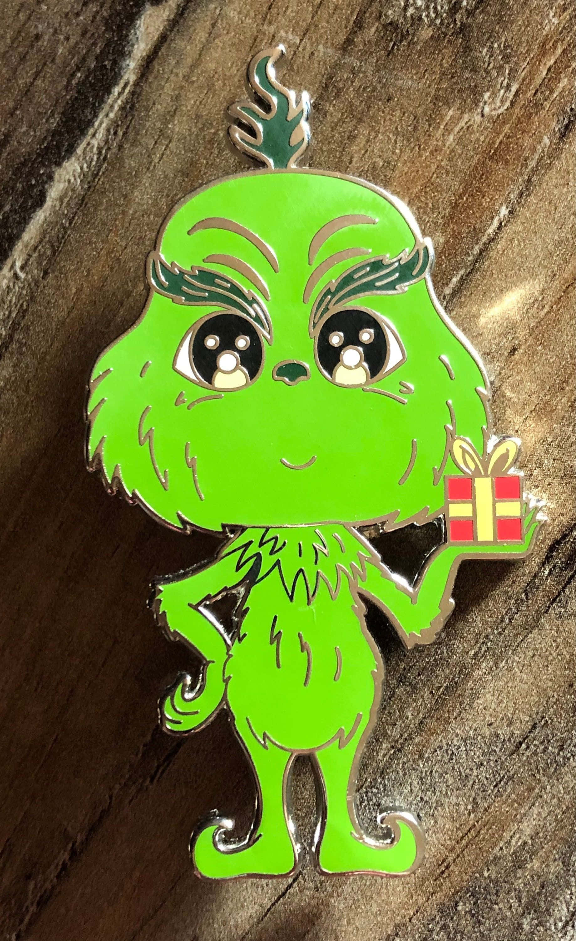 用這款可愛的格林奇散播聖誕精神！ 壁紙 — 一個可愛的Q版綠色鬼靈精琺瑯別針拿著禮物，放在質樸的木質表面上。