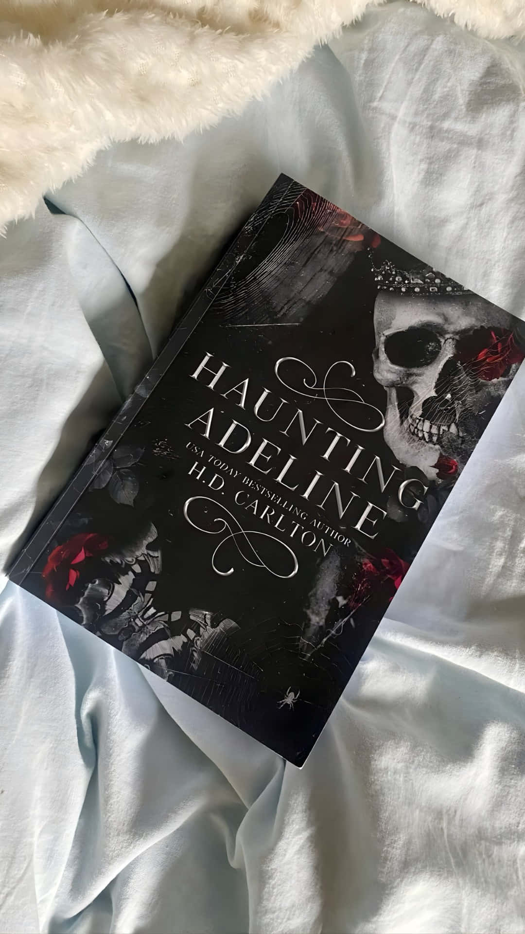  壁紙 — 一本名為《Haunting Adeline》的暗黑哥德式書籍，放在柔軟的白色床罩上，旁邊是一條蓬鬆的白色毛毯。