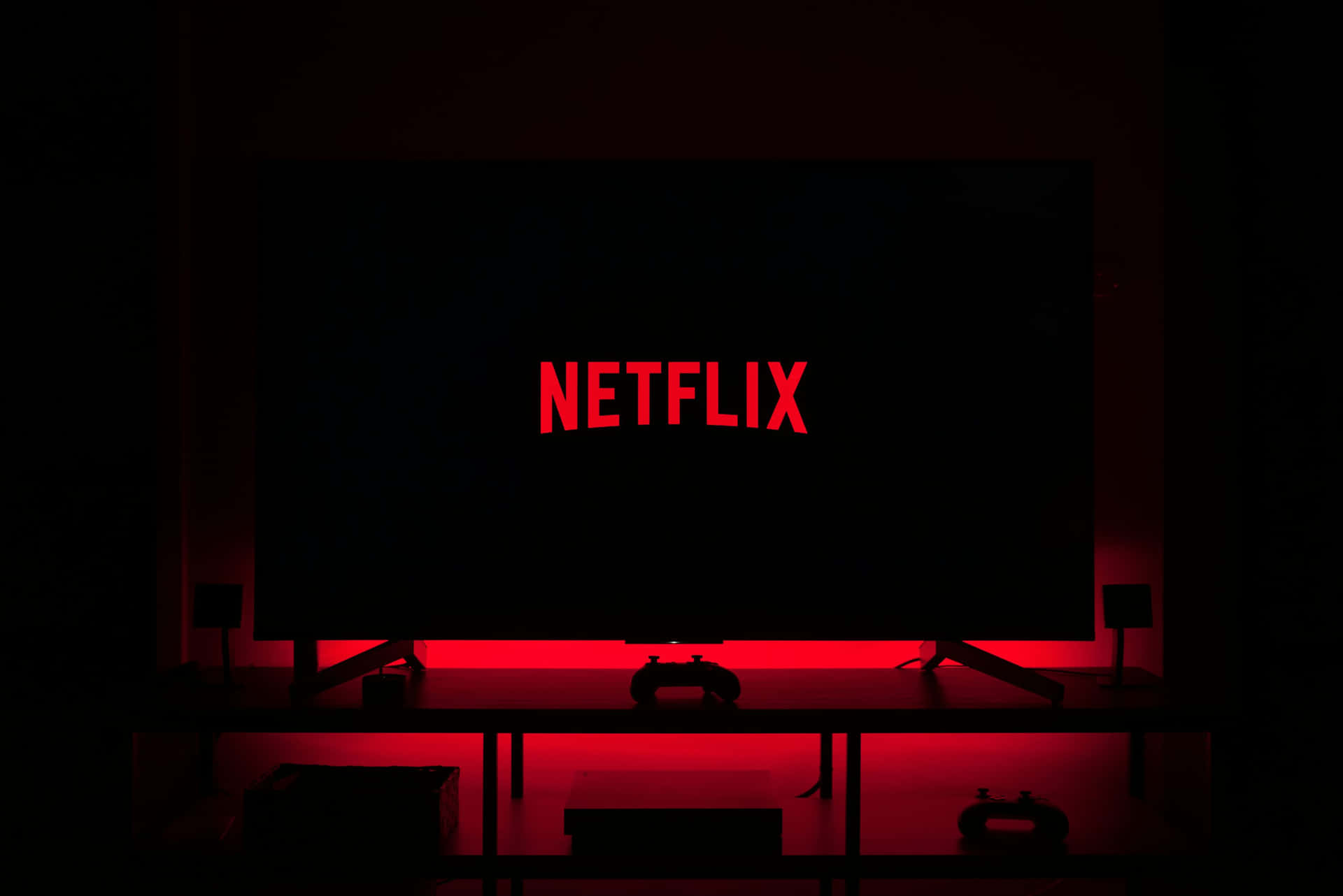  壁紙 — 一個黑暗的房間，被電視屏幕上發光的紅色Netflix標誌和下方的遊戲控制器照亮。