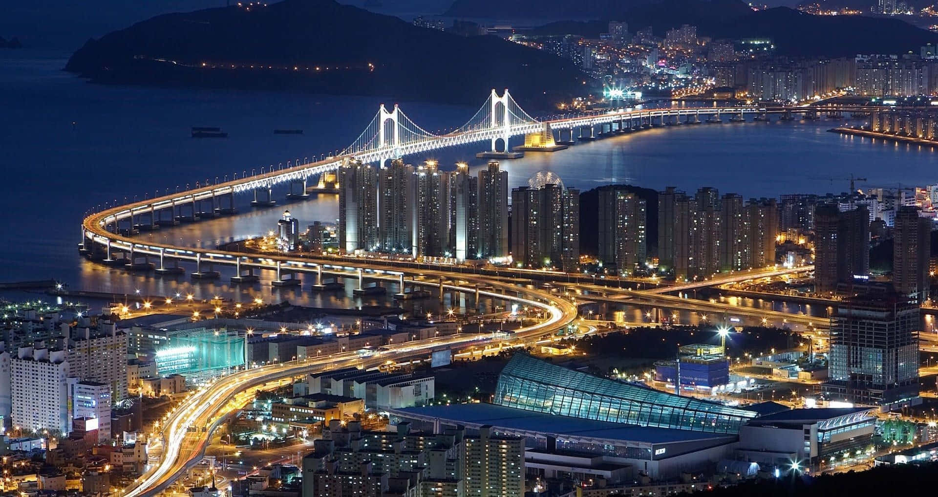 夜晚海灣上的一座發光吊橋，伴隨著燈火通明的城市建築和高速公路。