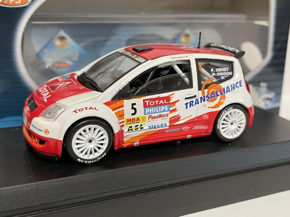 Citroen C2 Super 1600 桌布 壁紙 — 一個詳細的紅白拉力賽車模型，帶有贊助商貼紙，放置在透明盒內的黑色展示架上。