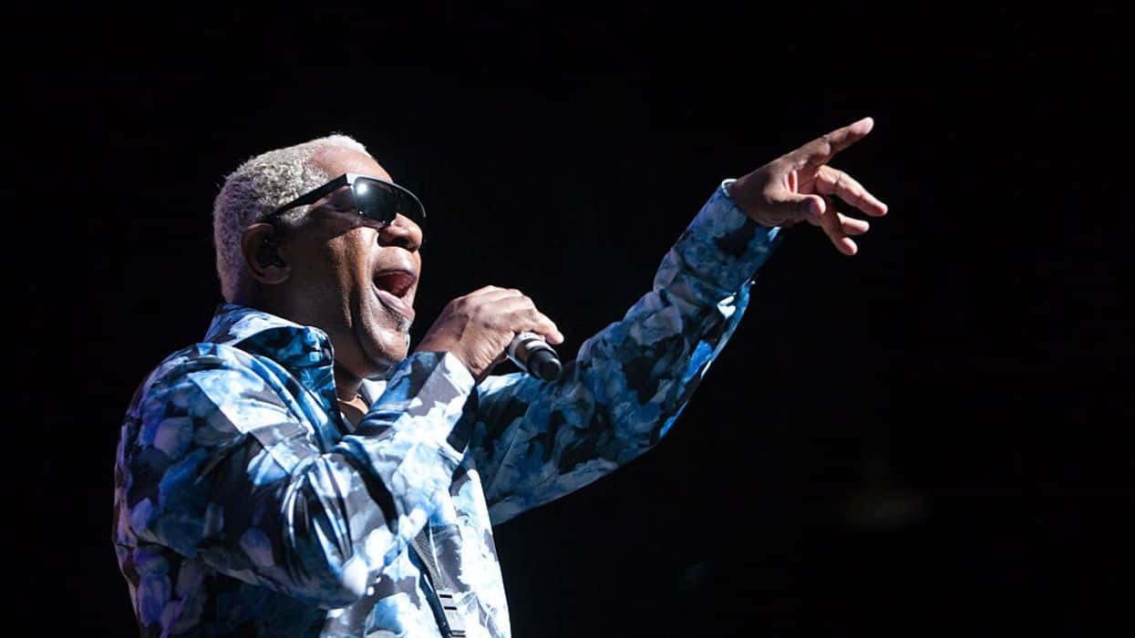  壁紙 — 一位身穿藍色圖案外套和太陽眼鏡的動感表演者，在黑暗的舞台背景下，一邊熱情地對著咪高峰唱歌，一邊向前指。