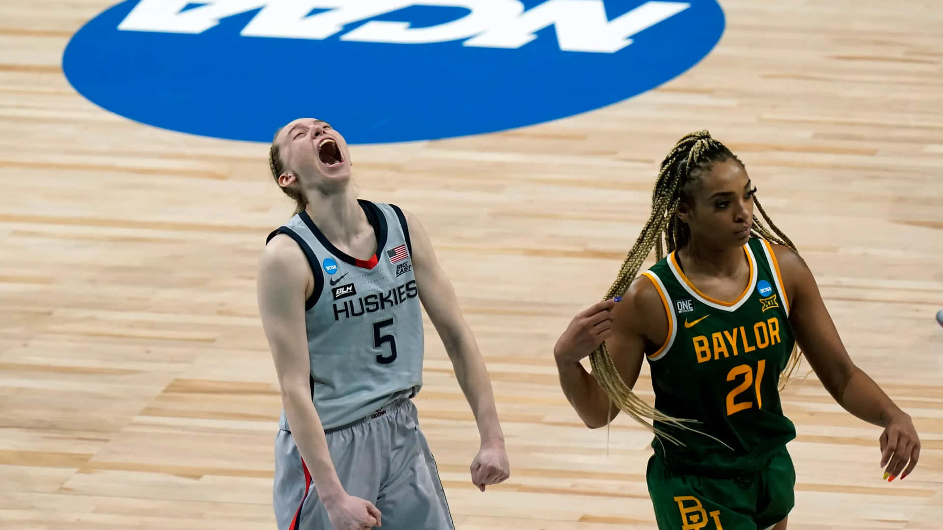  壁紙 — 一位身穿灰色Huskies球衣的女籃球員在木地板上勝利地尖叫，而一位Baylor球員走過。