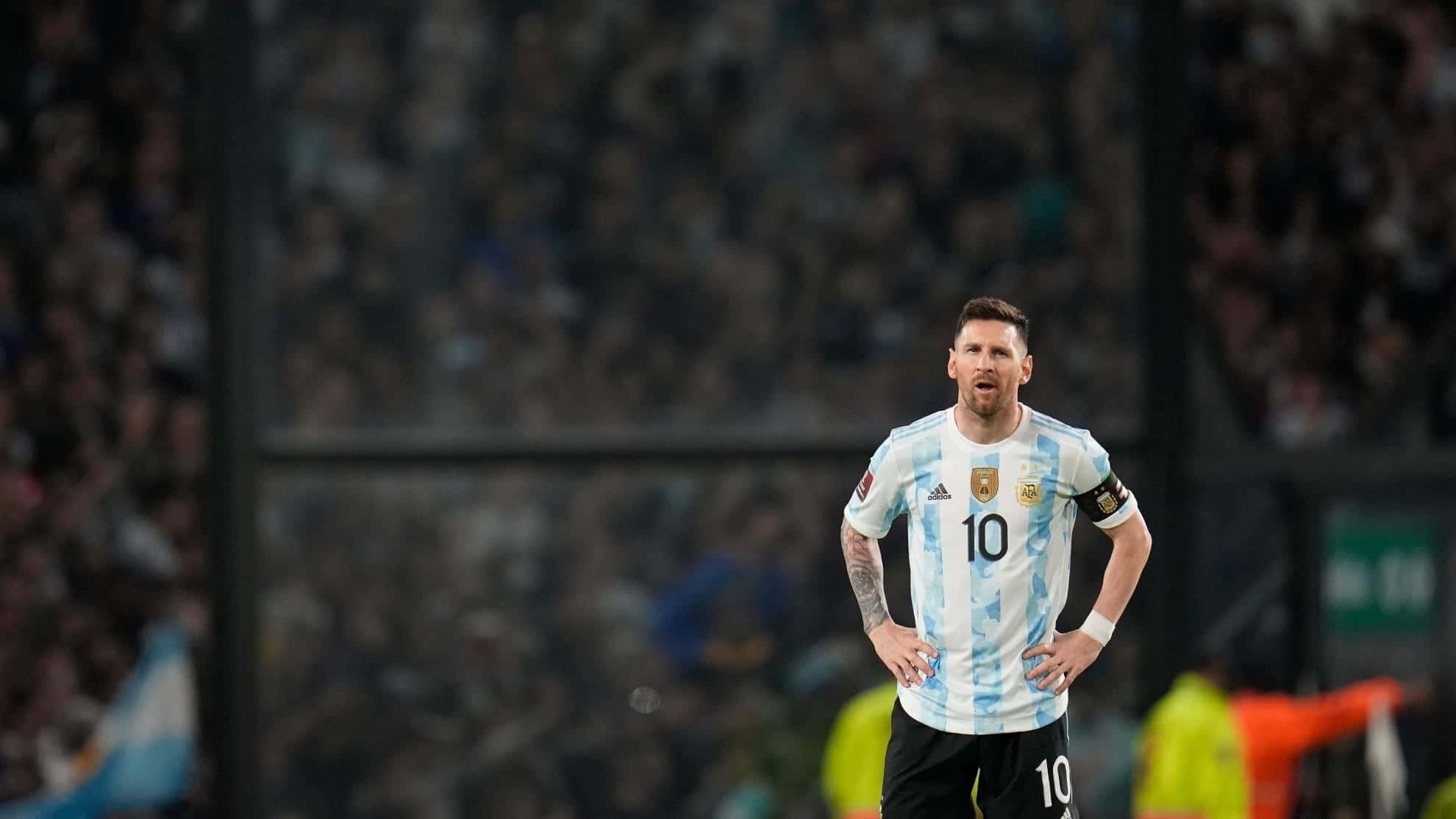 Messi 2022 桌布 壁紙 — 阿根廷足球巨星美斯身穿藍白間條球衣，在明亮的燈光下雙手叉腰站在球場上。