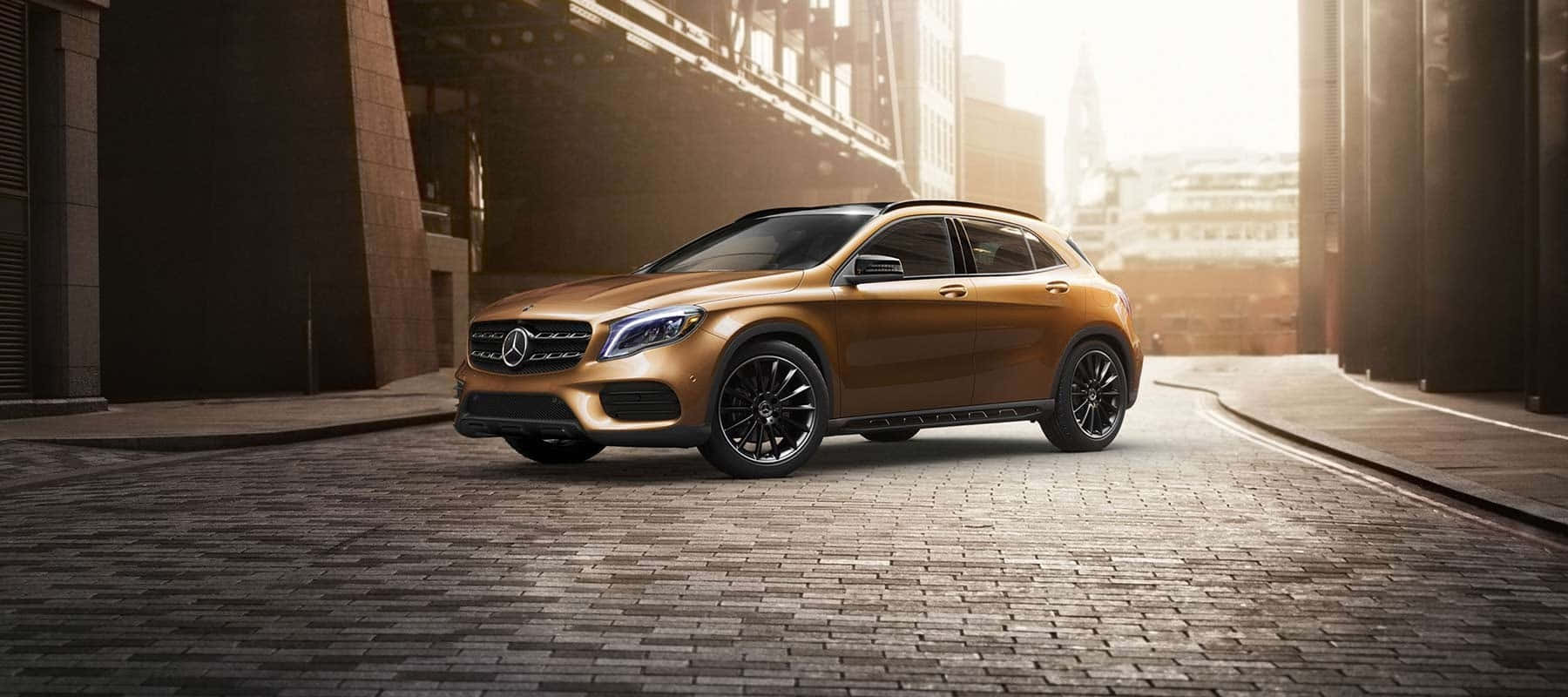 Mercedes Benz Gla-class 桌布 壁紙 — 一輛閃閃發光的金色 Mercedes-Benz GLA-Class SUV 停泊在現代建築之間陽光普照的鵝卵石小巷中。