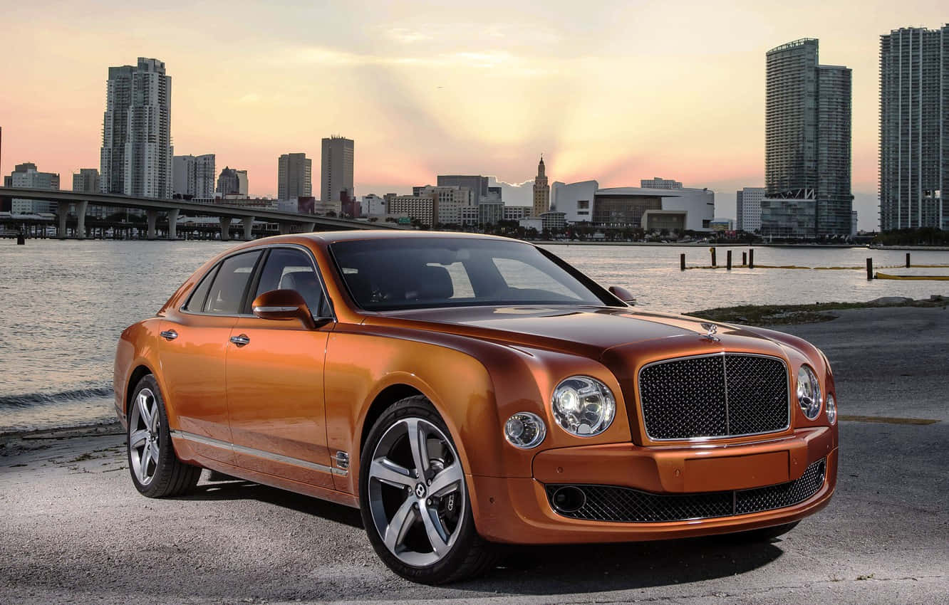Bentley Mulsanne 桌布 壁紙 — 一輛閃亮的橙色賓利轎車停在水邊，城市天際線在日落時分閃耀。