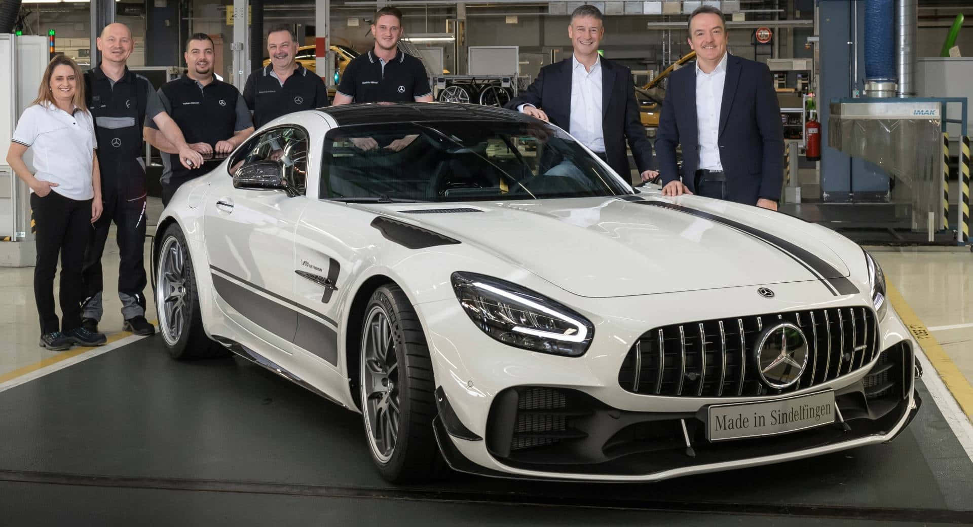  壁紙 — 一輛閃亮的白色Mercedes-AMG GT S跑車帶有黑色賽車條紋，在工廠環境中展示，周圍是穿著品牌服裝的微笑員工。