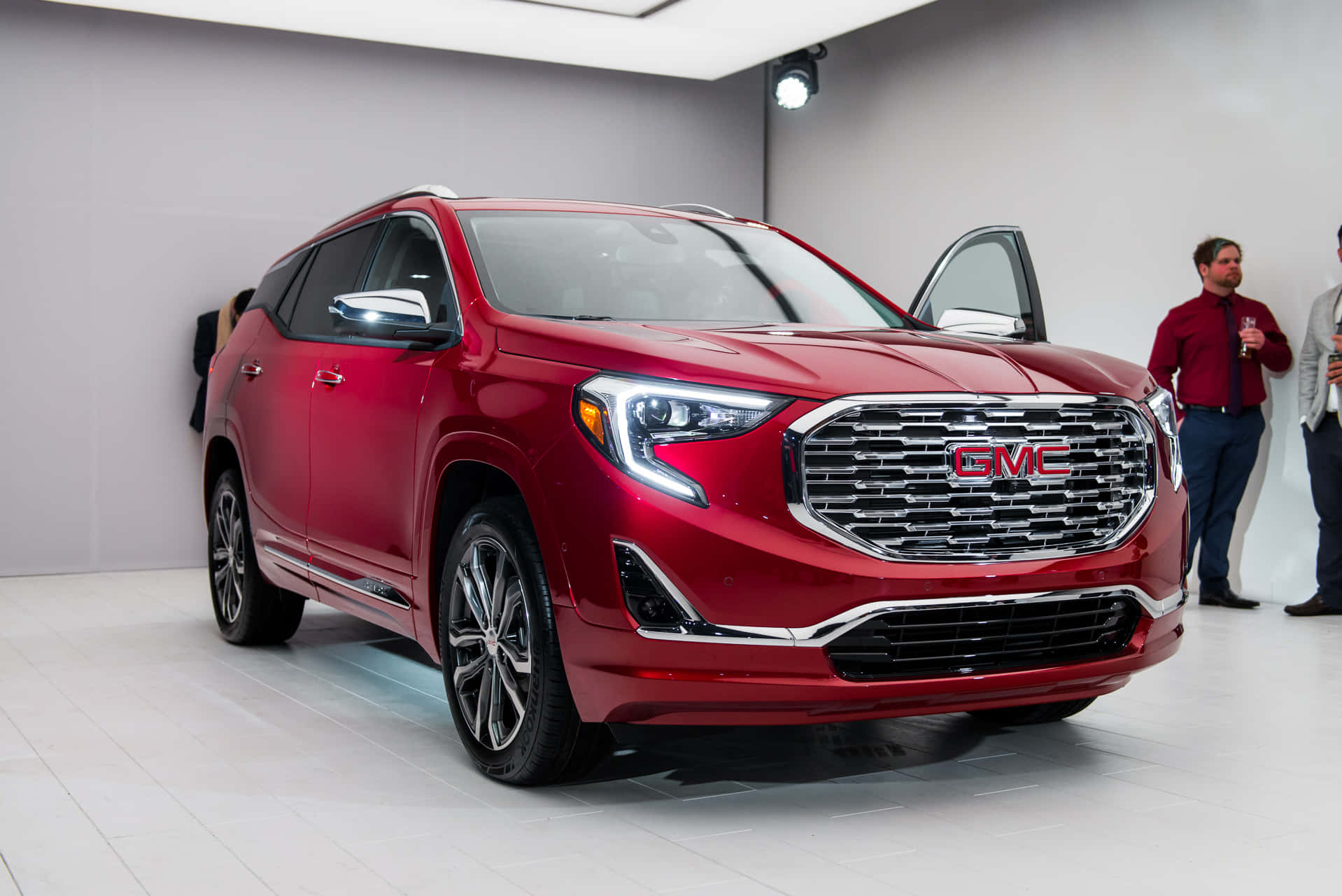 Gmc Terrain 桌布 壁紙 — 一輛光亮的紅色GMC Acadia SUV在明亮的室內車展中展示，司機門打開，附近有人觀察。