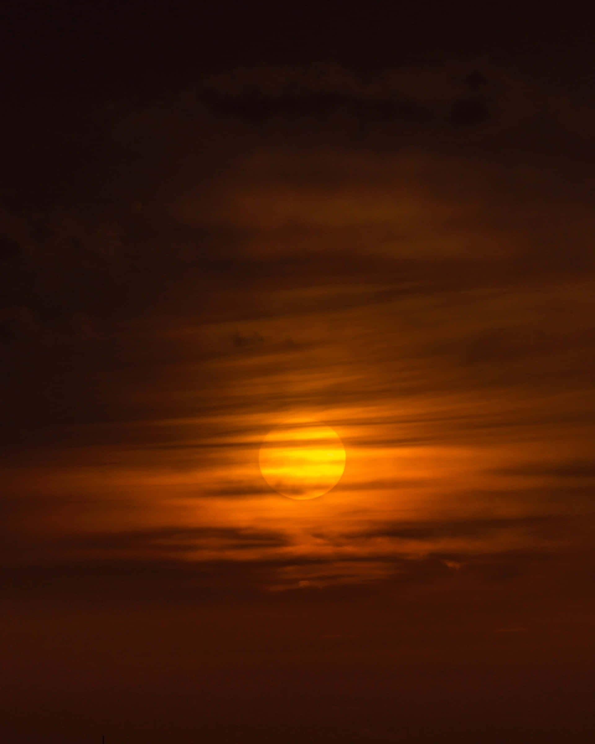 Sunset Hues 桌布 壁紙 — 一顆發光的橙色太陽在條紋雲後落下，將溫暖的光線投射在漸暗的天空上。