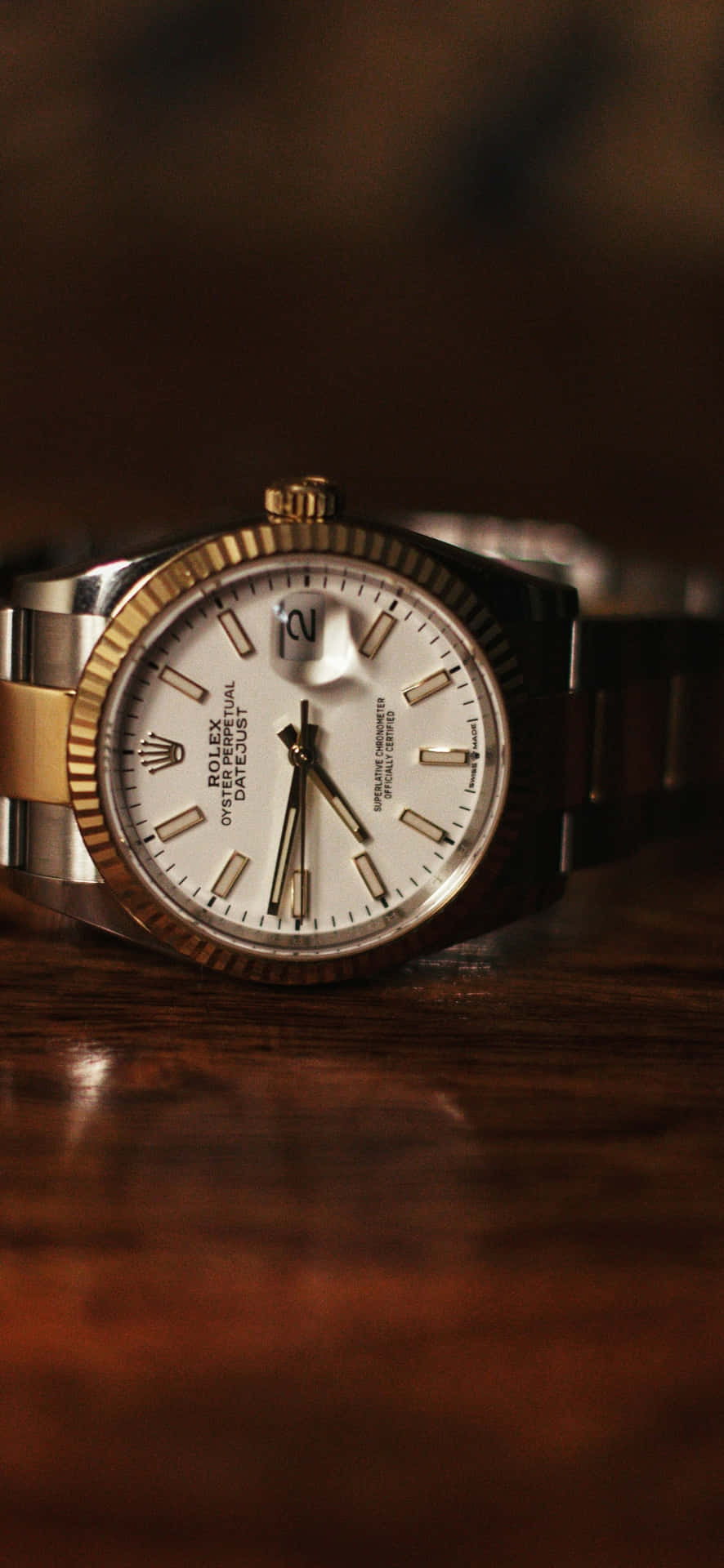  壁紙 — 一隻金色和鋼製的勞力士 Datejust 手錶優雅地放置在溫暖環境照明下的豐富木質表面上。