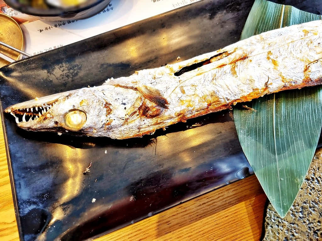  壁紙 — 一條烤魚，有尖銳的牙齒和金棕色魚皮，放在黑色碟子上，旁邊有一片綠葉。
