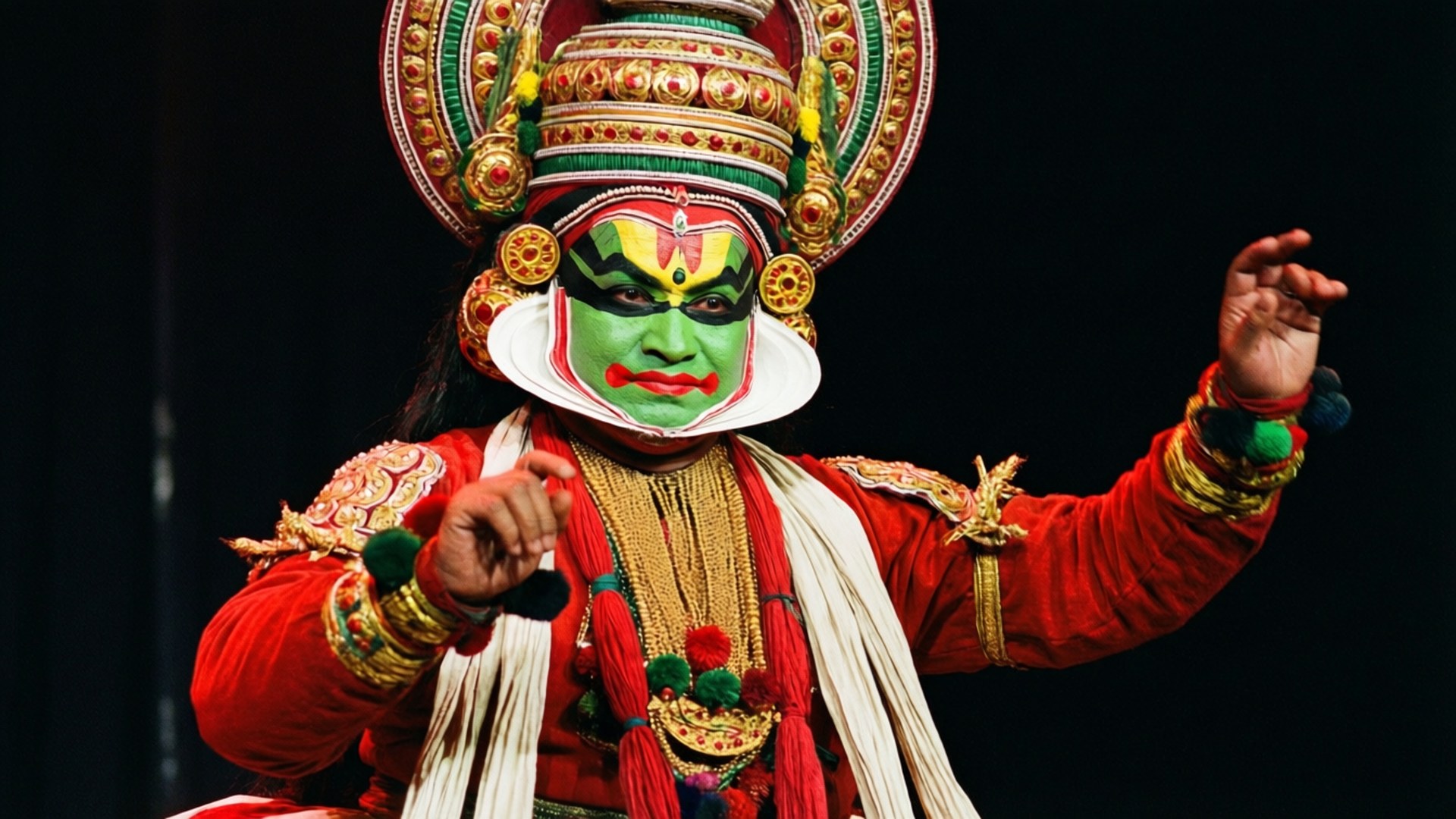 Onam 桌布 壁紙 — 一位卡塔卡利舞者，臉上塗有鮮豔的綠色顏料，身穿華麗的紅色服裝，在黑暗的舞台上以富有表現力的手勢表演。