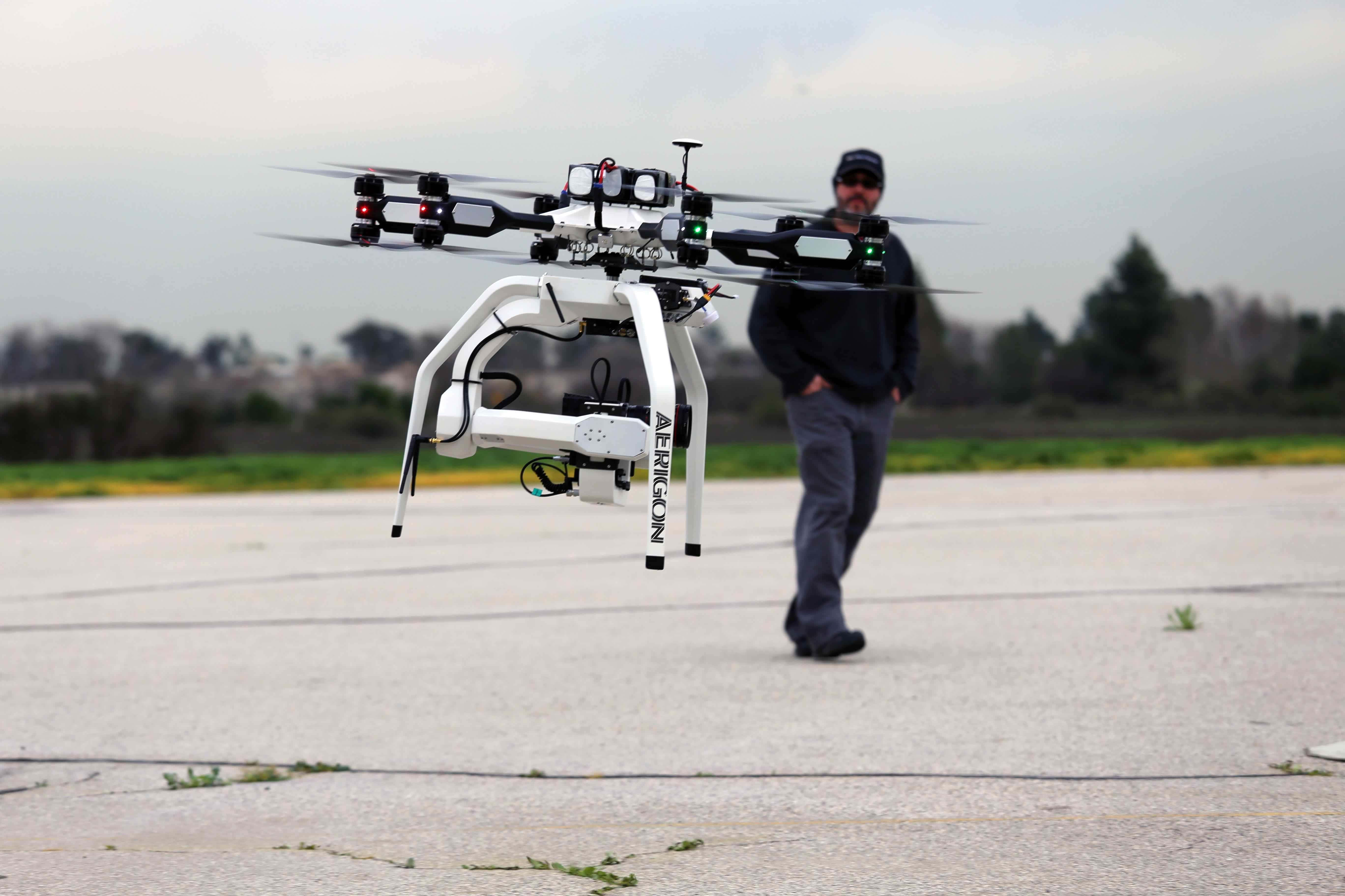 Drone Camera 桌布 壁紙 — 一架大型白色ArduPilot無人機在鋪砌的停車場上空盤旋，一名戴帽子的男子在它後面走過。