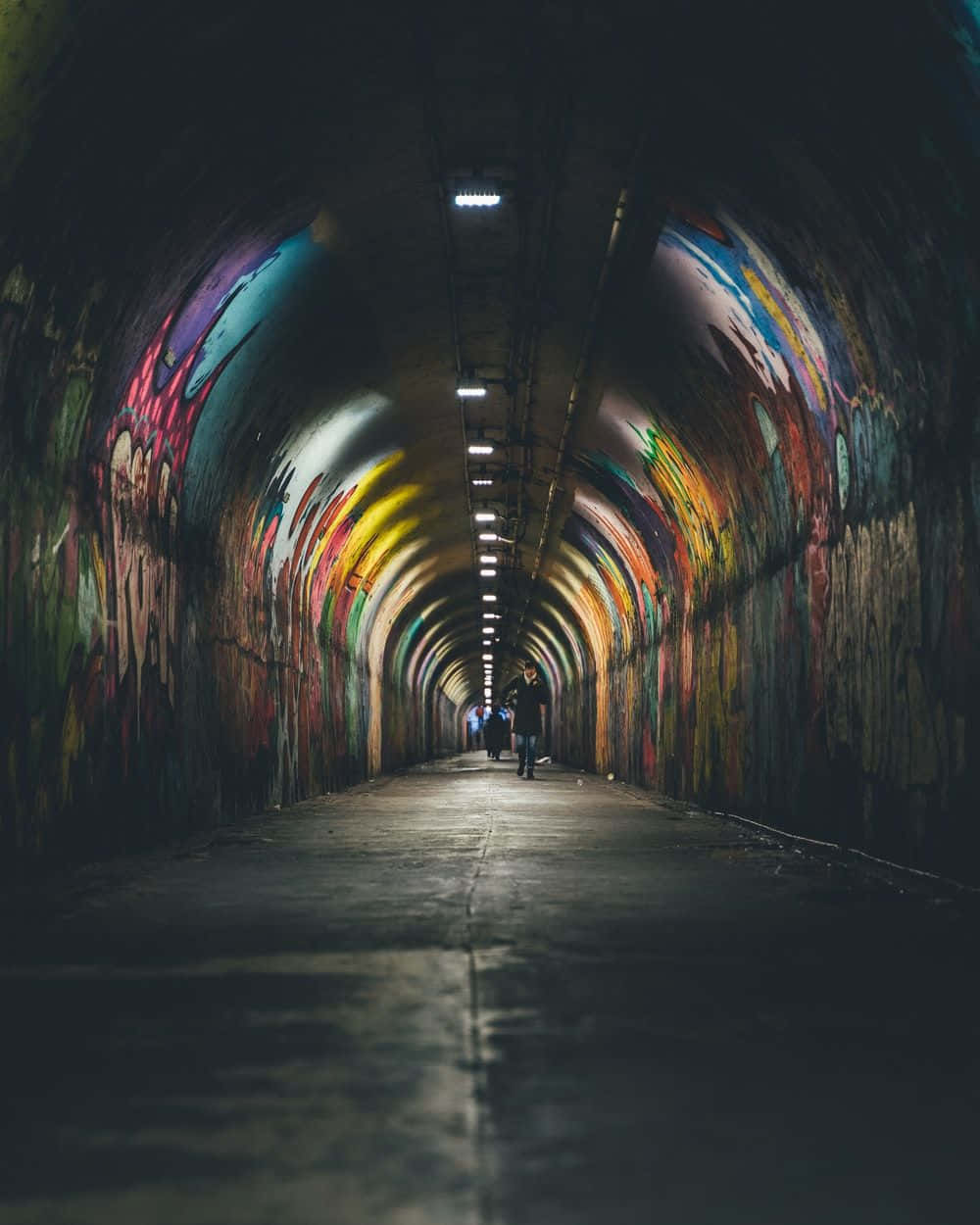 壁紙 — 一個孤獨的身影走過一條燈光昏暗的城市隧道，隧道彎曲的牆壁上裝飾著充滿活力的塗鴉藝術。