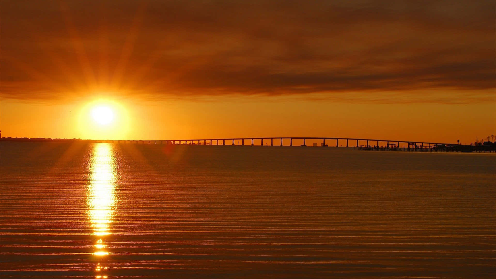 Sunset Reflection 桌布 壁紙 — 一條長橋橫跨平靜的水面，夕陽如火般橙紅，在水面上投射出一條燦爛的路徑。