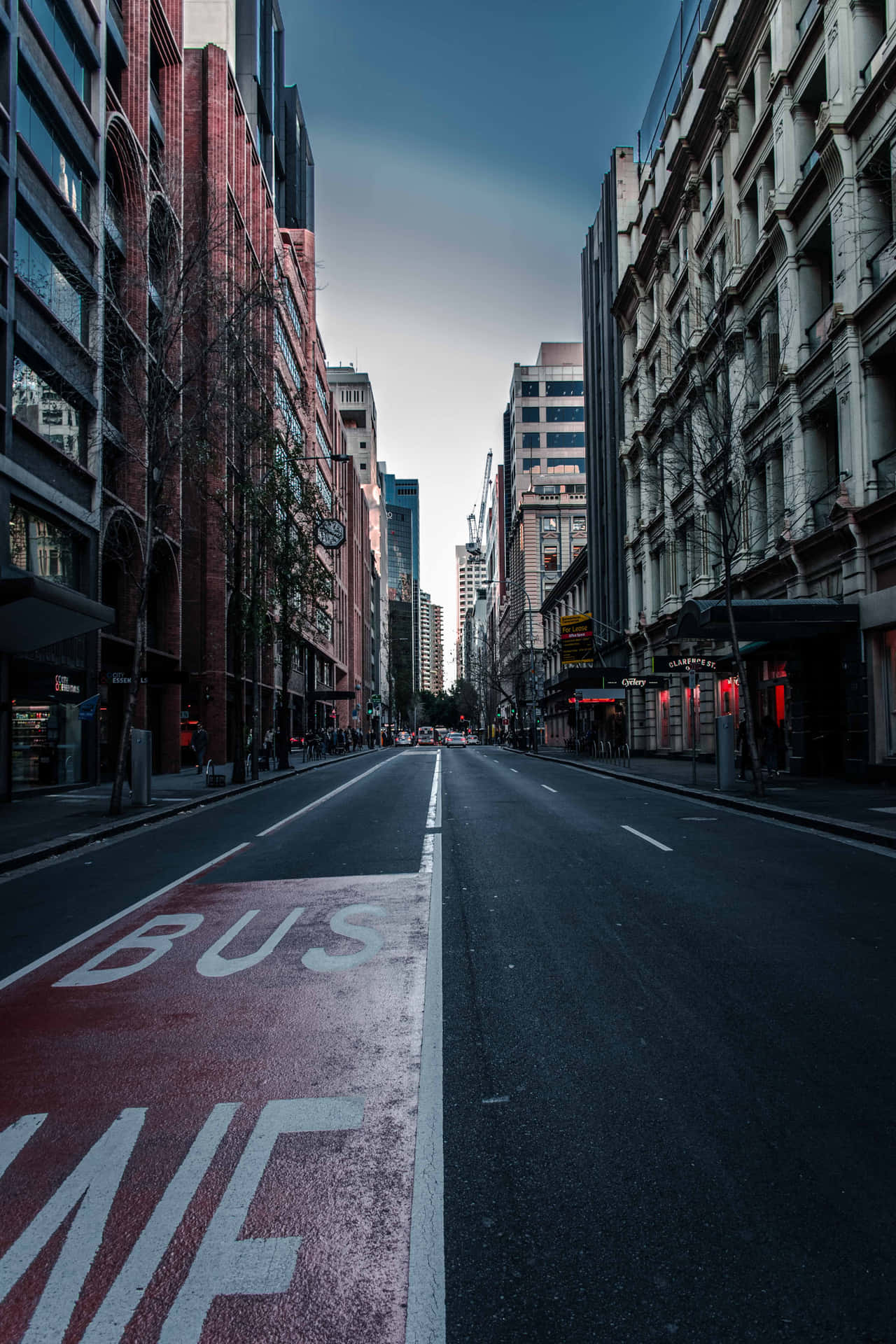  壁紙 — 一條長長的空蕩蕩的城市街道，兩旁是高樓大廈，在陰鬱的暮色天空下，帶有紅色巴士專用道標記。
