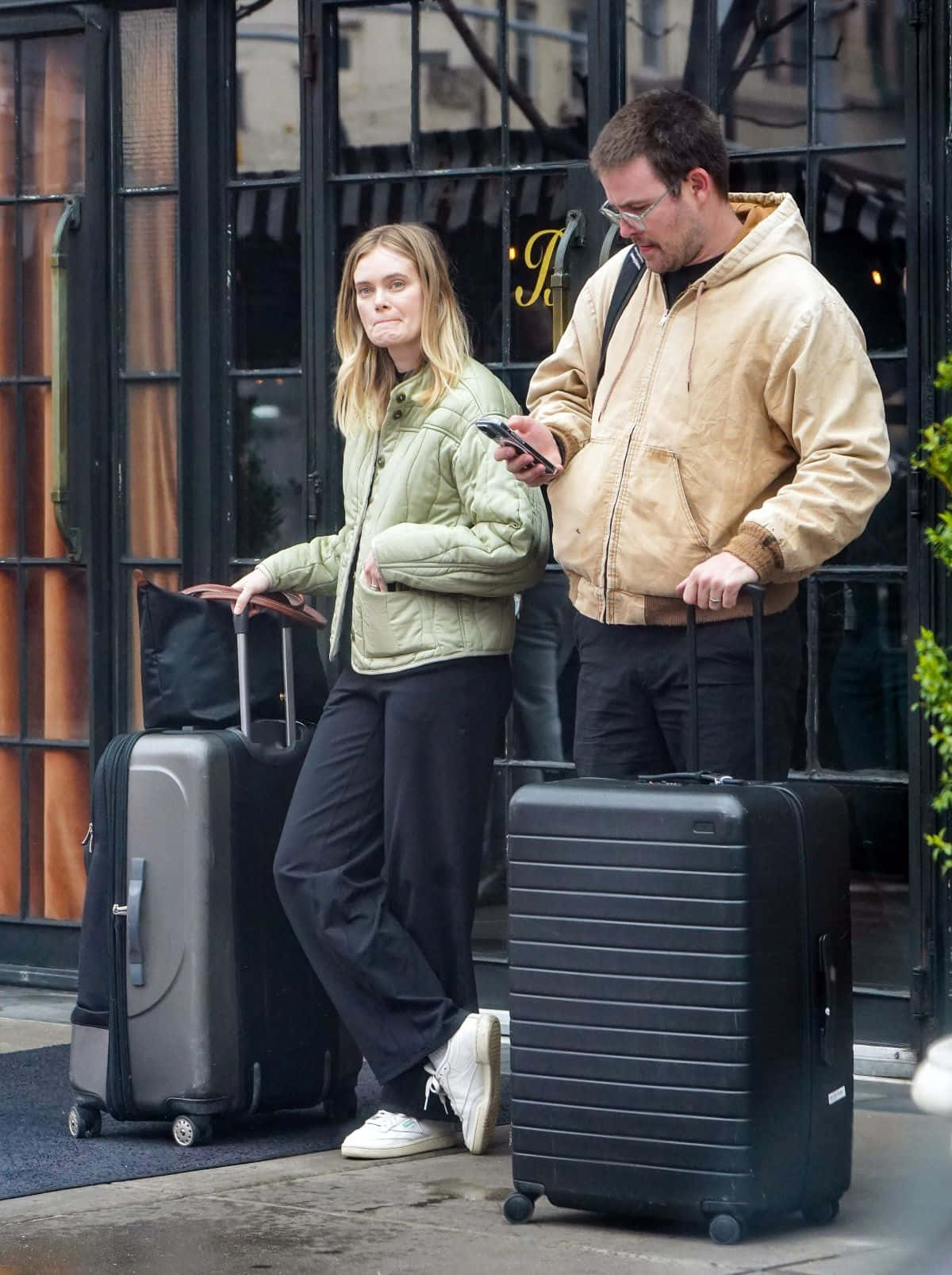 Sara Paxton 桌布 壁紙 — 一男一女站在建築物外，拖著滾輪行李箱，一人查看手機，另一人向前看