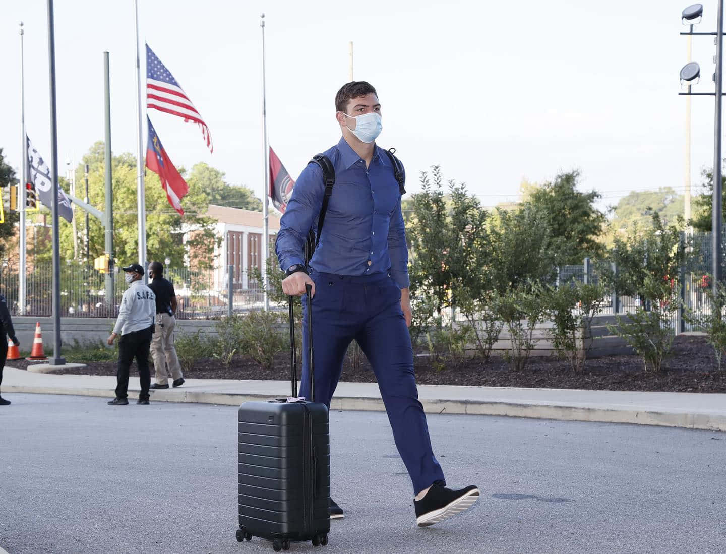  壁紙 — 一名身穿藍色襯衫和口罩的男子自信地走過鋪砌區域，拖著黑色行李箱，旗幟下半旗。