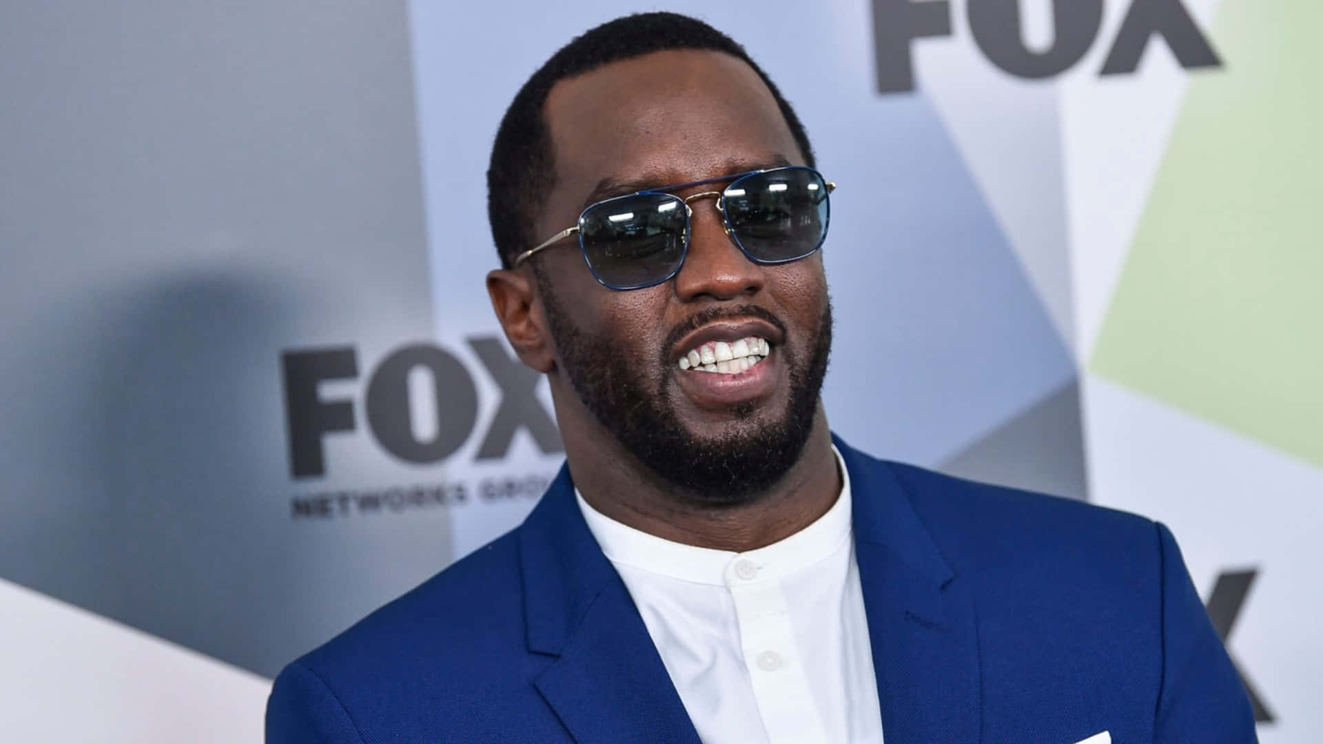  壁紙 — 一名身穿藍色西裝和太陽眼鏡的男子在FOX Networks Group活動背景前燦爛微笑。
