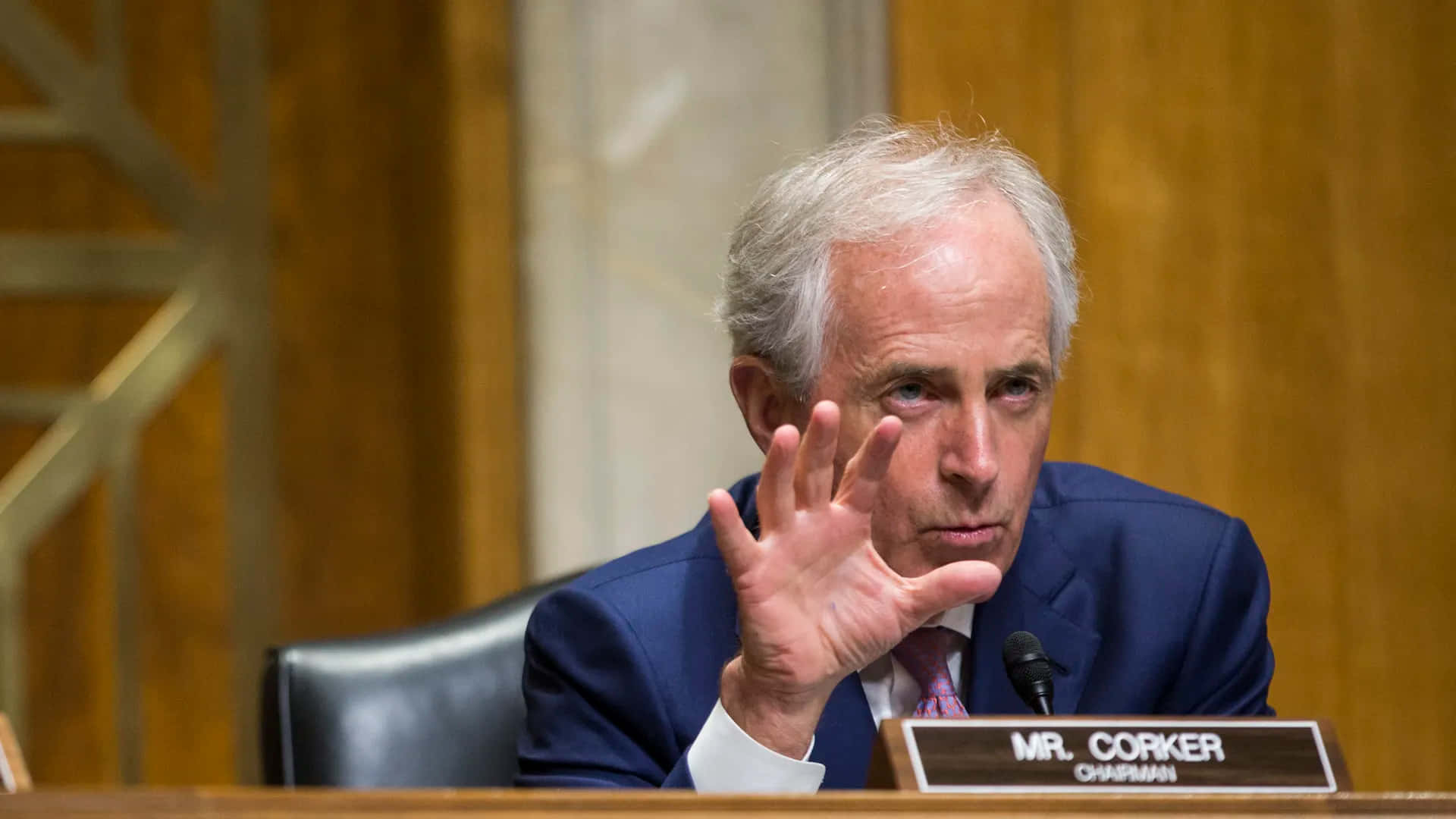  壁紙 — 一名身穿藍色西裝的男子坐在木桌前，桌上放著名牌寫著 MR CORKER CHAIRMAN，他正用手做手勢。