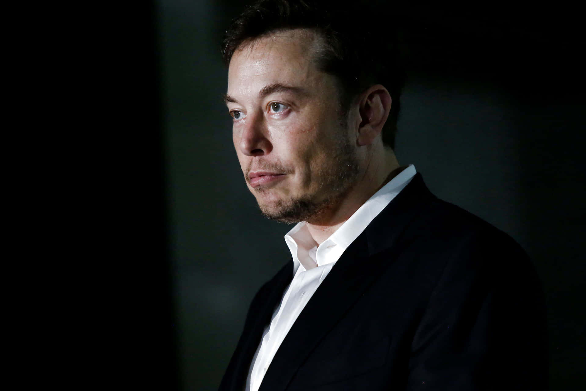 一個身穿深色西裝和白色襯衫的男人，在昏暗環境中若有所思地望向一旁。