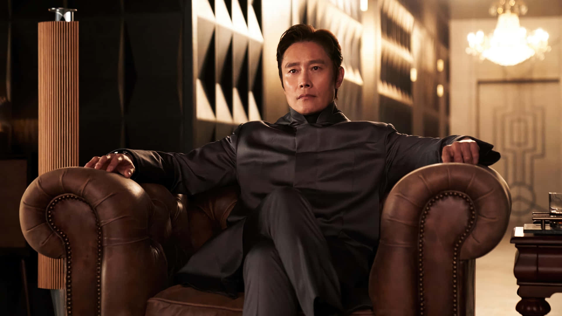  壁紙 — 一名身穿深色西裝的男子自信地坐在一張棕色皮扶手椅上，房間豪華，光線溫暖，配有幾何牆板和吊燈。