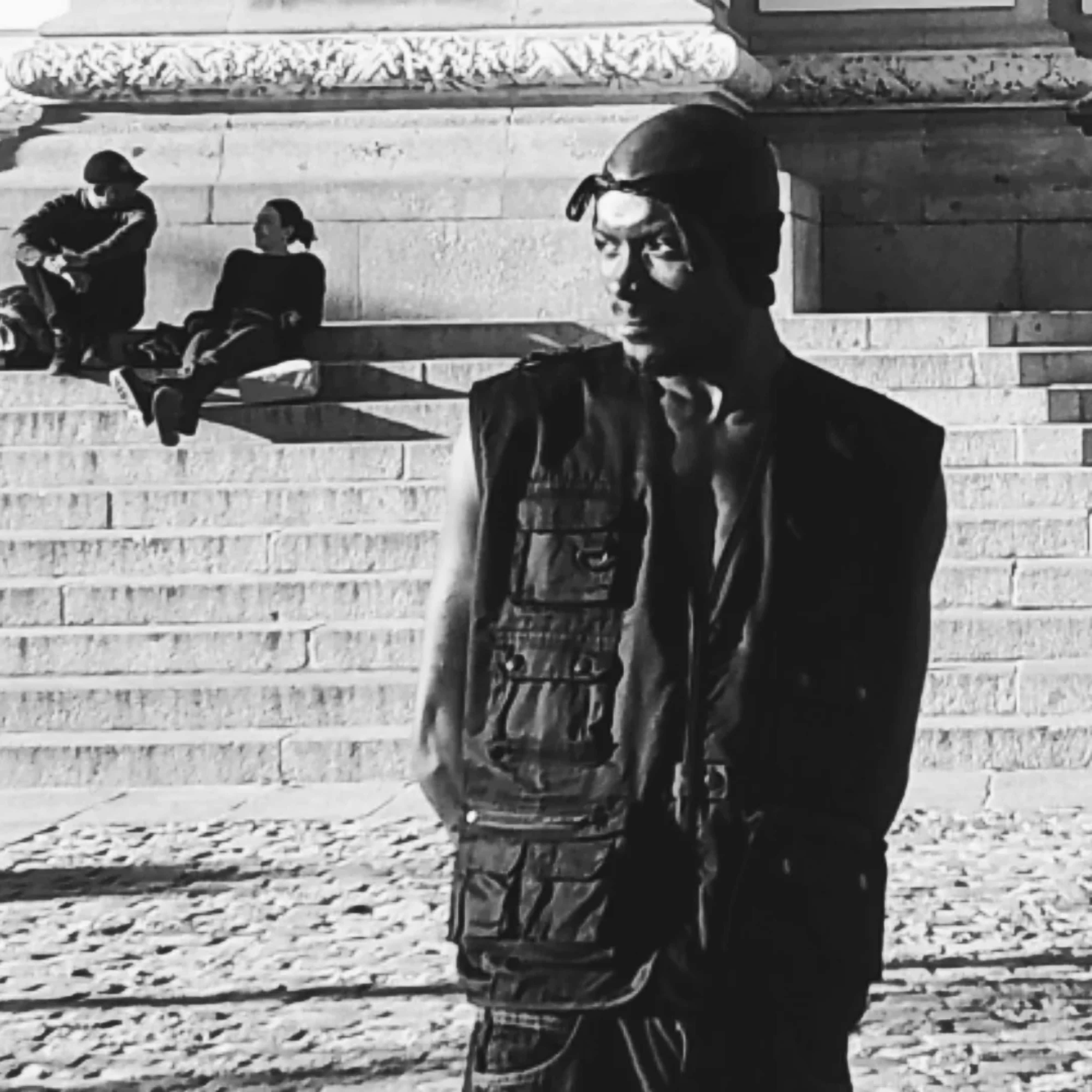 Cris Molo Akapo 壁紙 — 一個穿著背心和帽子的男人以鮮明的黑白對比站立，身後有另外兩人坐在石階上。