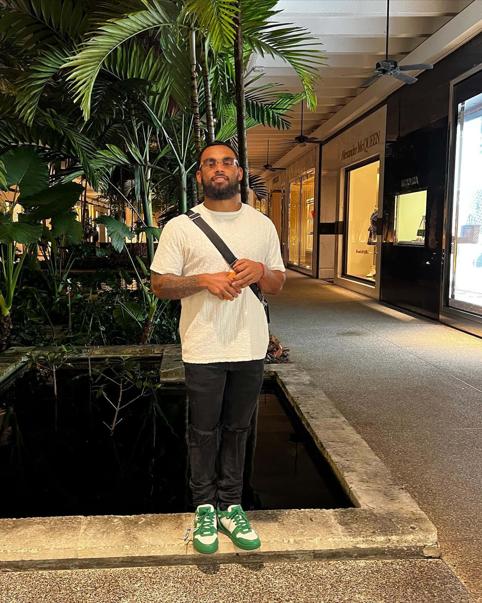  壁紙 — 一名身穿白色T恤和綠色運動鞋的男子自信地站在一個綠意盎然的室內購物拱廊的水景旁。
