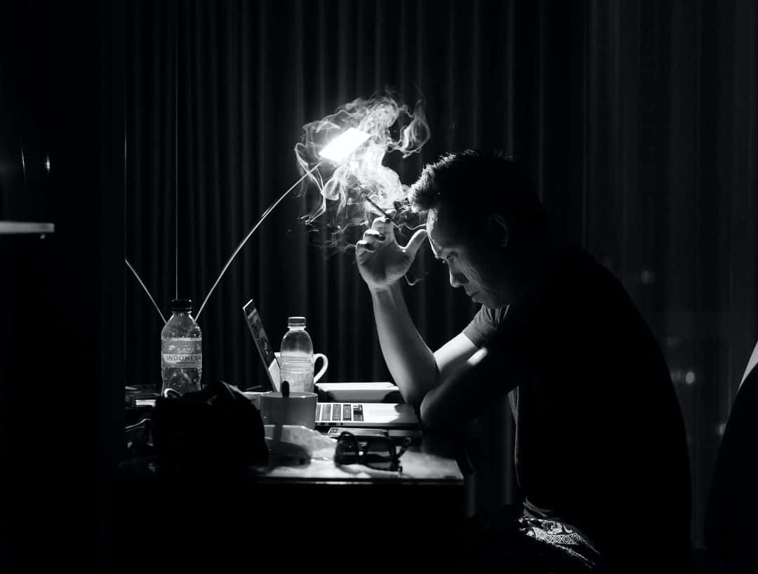 男人在燈光昏暗的書桌上吸煙，一盞燈投射出戲劇性的陰影。