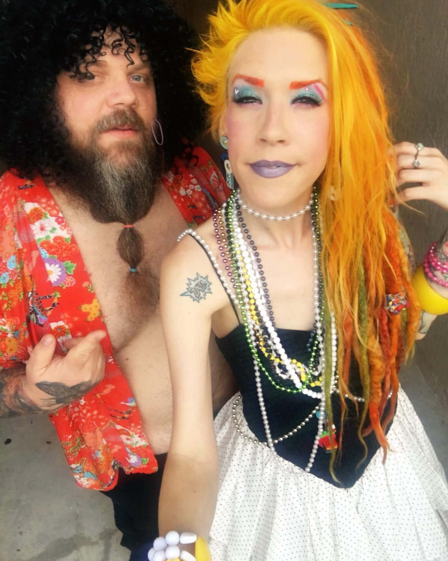  壁紙 — 一個戴著黑色捲曲假髮和鬍鬚的男人站在一個擁有鮮豔橙色頭髮和大膽妝容的女人旁邊，她穿著多層珠子和圓點連衣裙。