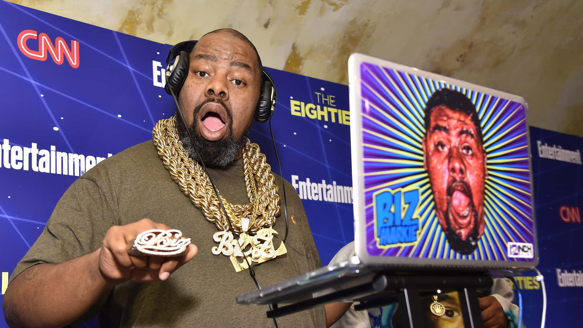  壁紙 — 一名戴著耳機和金鏈的男子手持「Biz Markie」曲奇，而手提電腦在充滿活力的太陽爆發背景上顯示他驚訝的表情。