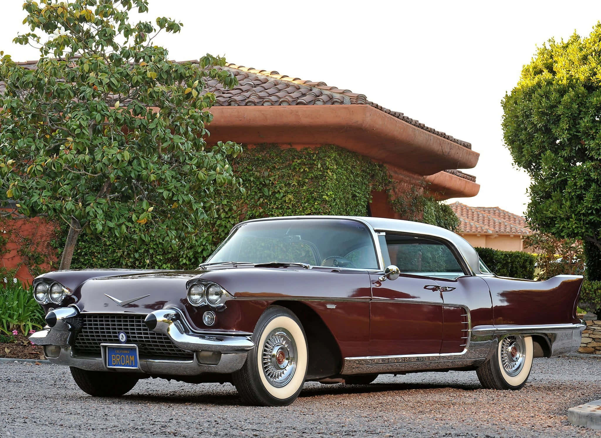 Cadillac Eldorado 桌布 壁紙 — 一輛栗色1959年凱迪拉克，帶有鍍鉻裝飾和白色側壁輪胎，停在有綠植的灰泥房屋前的礫石上。