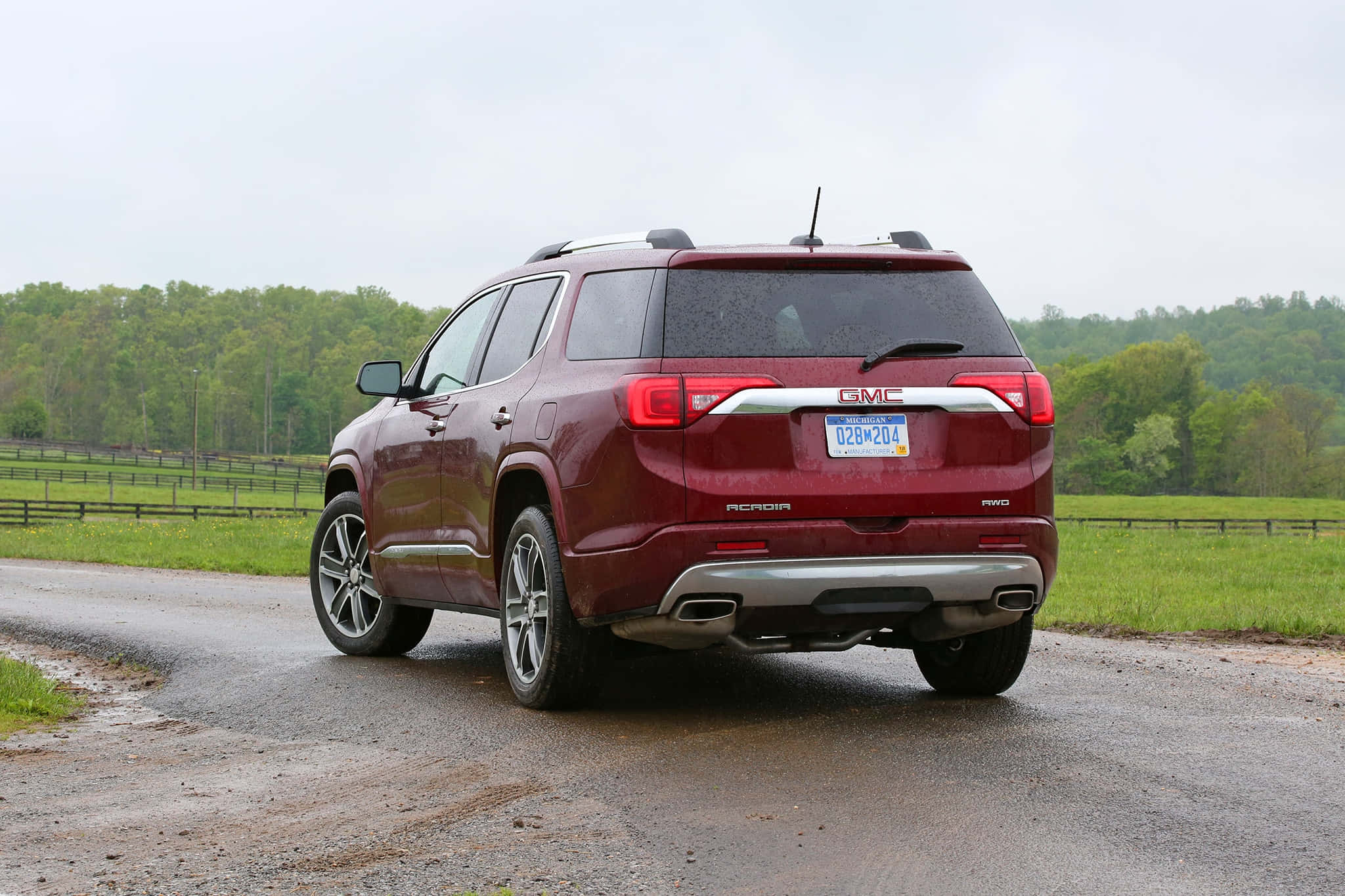 Gmc Acadia 桌布 壁紙 — 一輛栗色GMC Acadia SUV在陰天下行駛在濕潤的鄉村道路上，兩旁是綠色的田野和樹木。