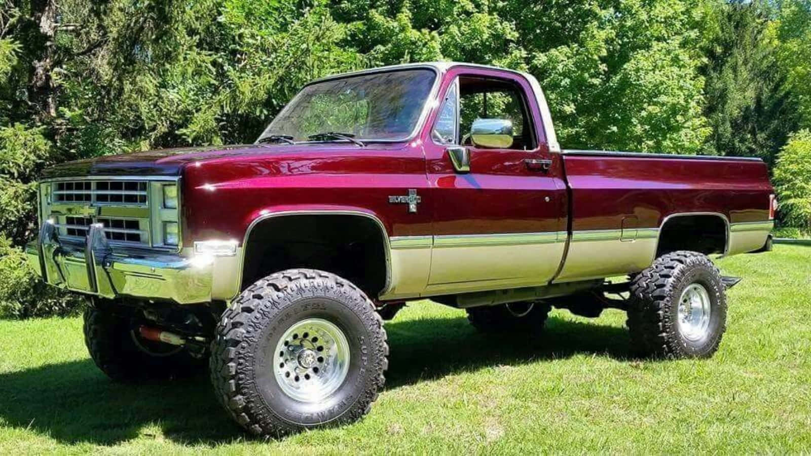 Square Body Truck 桌布 壁紙 — 一架栗色升高版雪佛蘭K10皮卡車，配備鍍鉻飾件和特大輪胎，驕傲地停泊在陽光明媚的草地上，四周環繞著茂密的綠樹。