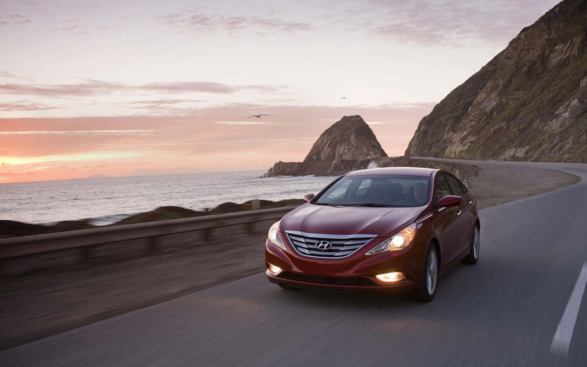 Hyundai Sonata 桌布 壁紙 — 一輛栗色轎車在日落時分沿著海岸公路飛馳，背景有海洋懸崖和岩石小島。