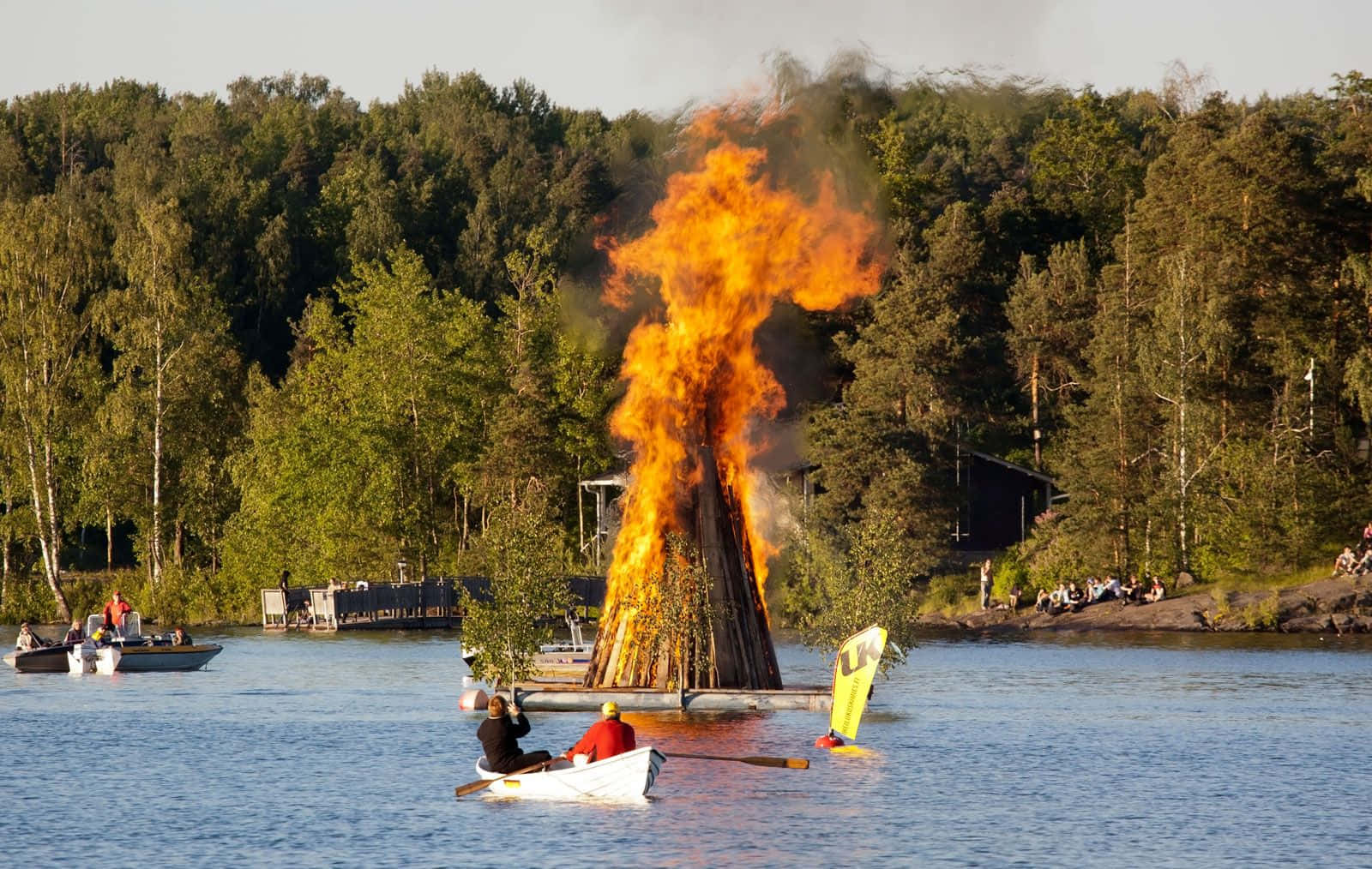  壁紙 — 一個巨大的篝火在湖上明亮地燃燒，人們在船上觀看並在附近划船，周圍是茂密的綠色森林。