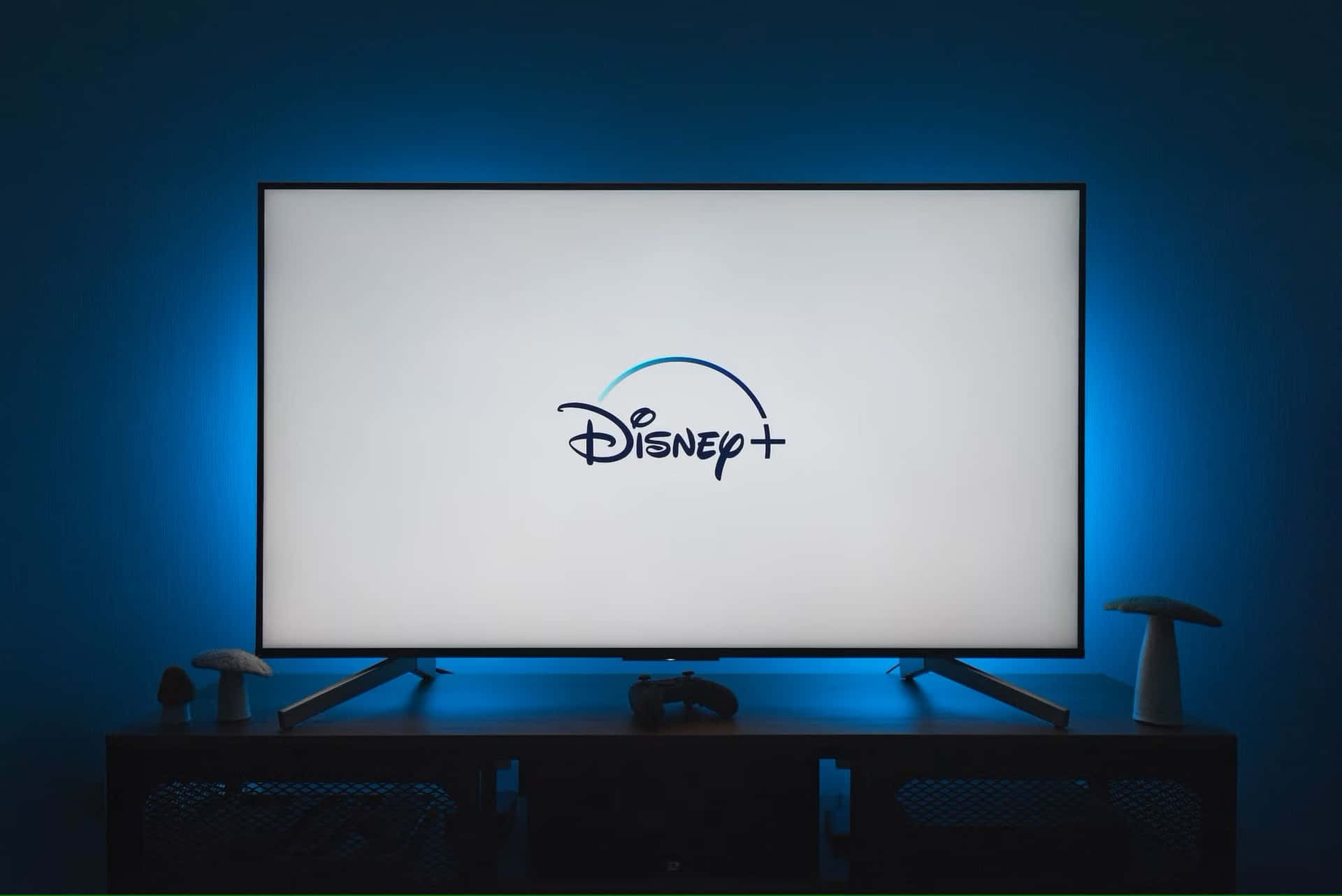  壁紙 — 一部現代電視在極簡娛樂設置中，顯示Disney+標誌，背景是發光的藍色環境光。