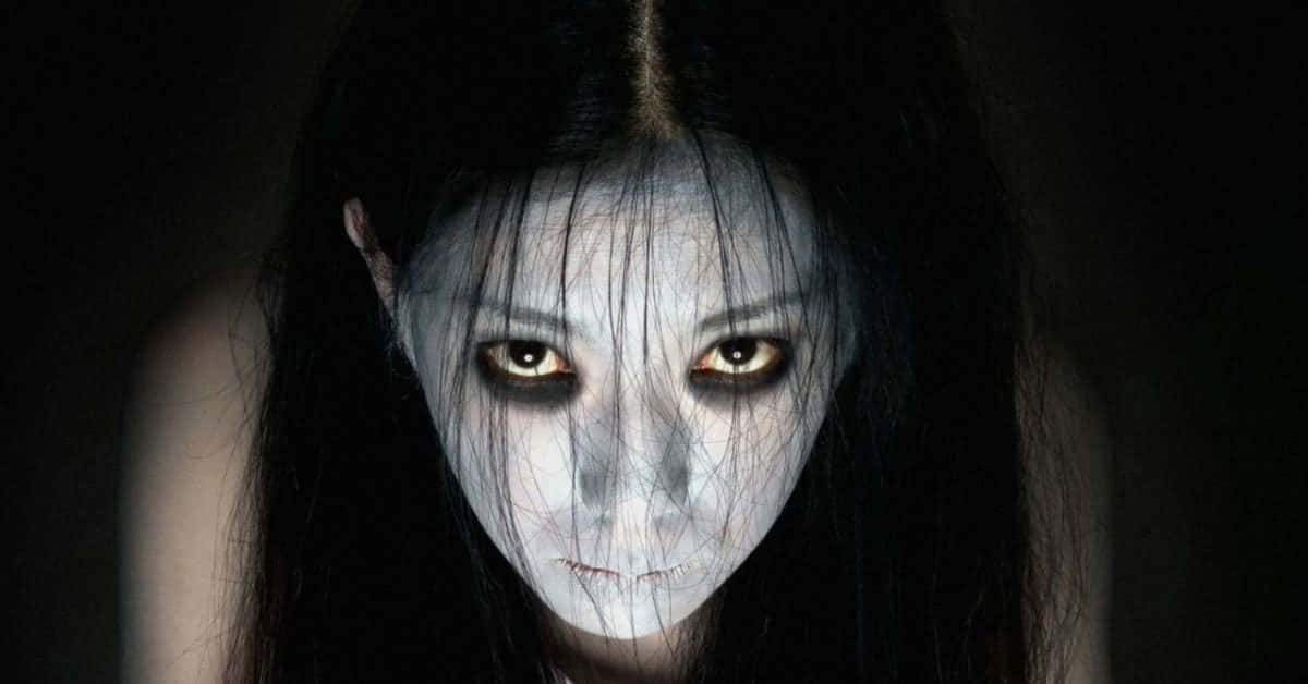  壁紙 — 一個蒼白幽靈般的女人，閃爍著琥珀色的眼睛，從黑暗中凝視，她的黑髮勾勒出一張詭異平靜的臉。
