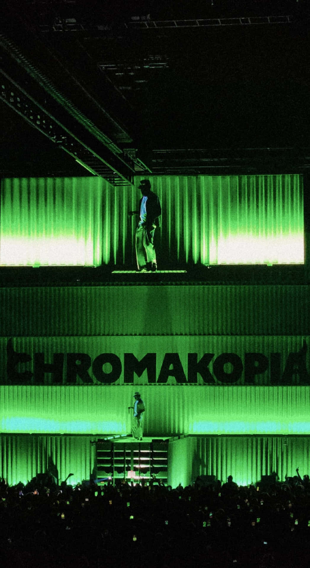 壁紙 — 一位表演者站在發光的綠色舞台上，下方亮著「CHROMAKOPIA」，在垂直光束中形成剪影，黑暗的人群中粉絲舉起手機。
