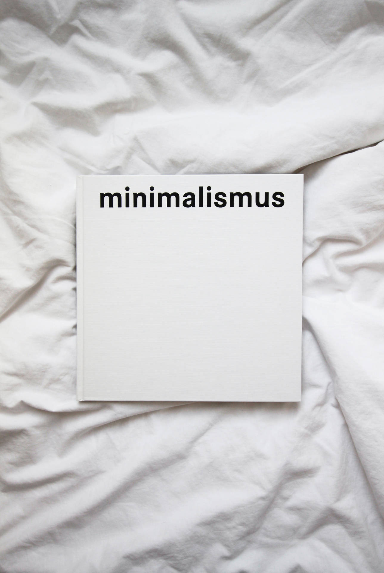極簡白色極簡主義字樣 壁紙 — 一本名為 minimalismus 的純白色書本，放在皺巴巴的白色床單上，構成極簡主義的構圖。
